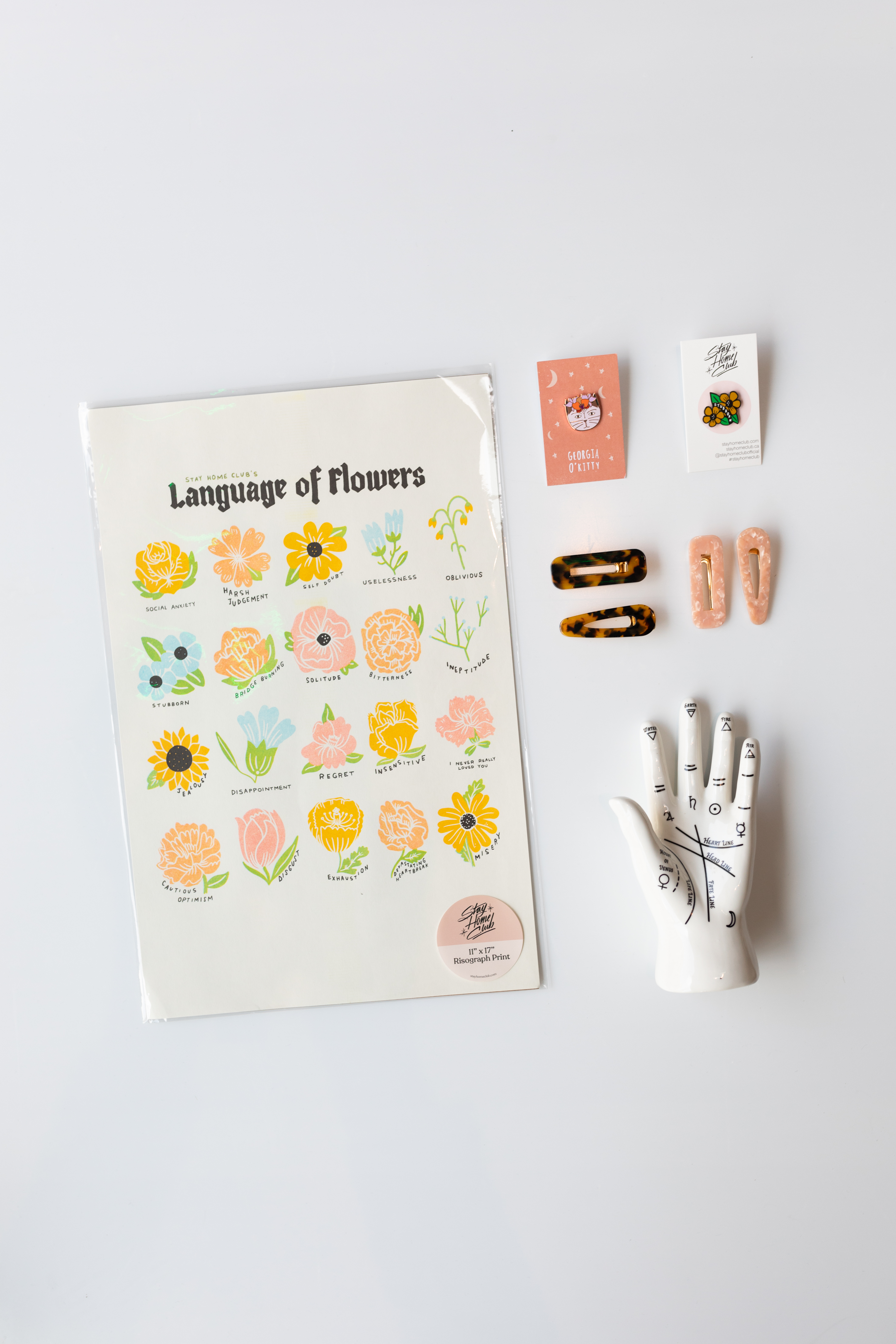 a print titled Language of Flowers depicting several drawings of pink, ﻿orange, blue, and yellow flowers, a pin of a cat with a flower crown titled Georgia O'Kitty; a pin of two flowers with the text reading stubborn; two tortise shell berets; two pink berets; a hand sculpture depicting handlines. 