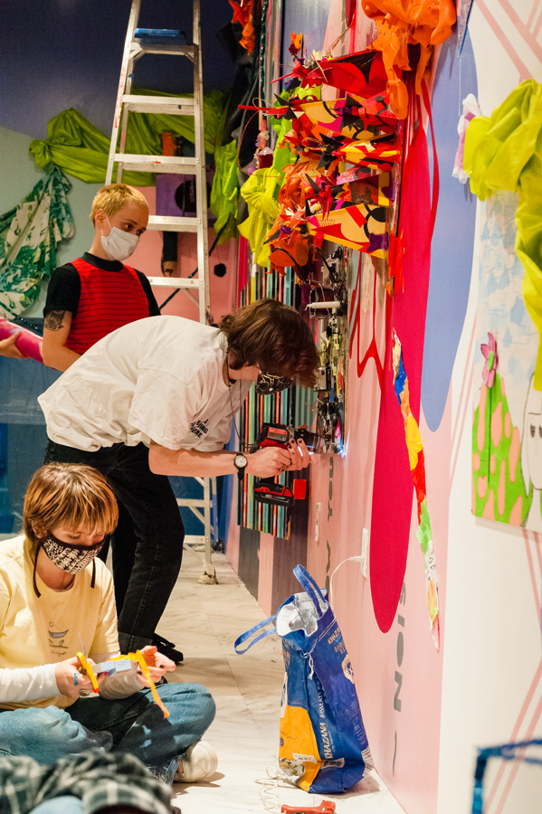 Close up image of a brightly colored large scale, 3D collage installation, that displays a wide range of thrifted materials. There are three people in the image working with their hands and working on the installation. 