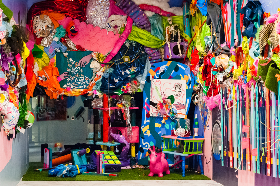 A brightly colored large scale, 3D collage installation, that displays a wide range of materials: a picket fence, cloth draped from and tied to the wall, large tube-like structures, a skeleton in the corner, a small pink unicorn on the floor, and other thrifted materials.