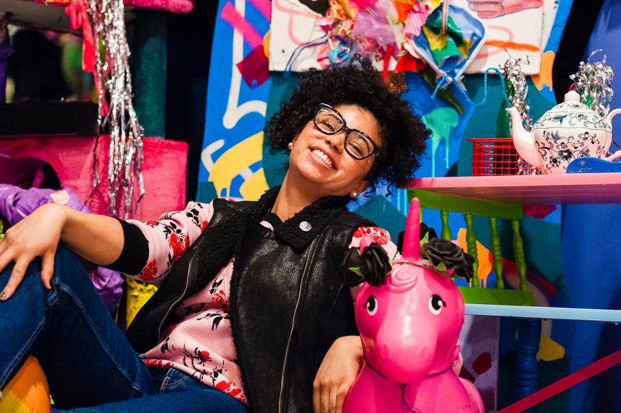 Artist Moe Gram sitting in front of brightly colored large scale, 3D collage installation, that displays a wide range of thrifted materials. She is leaning back on a pink blow up unicorn and is wearing a pink long sleeve, black vest, blue jeans, glasses with black frames, and her black curly hair is pulled up in a high ponytail. She has a big smile on her face and her head is cocked back. 
