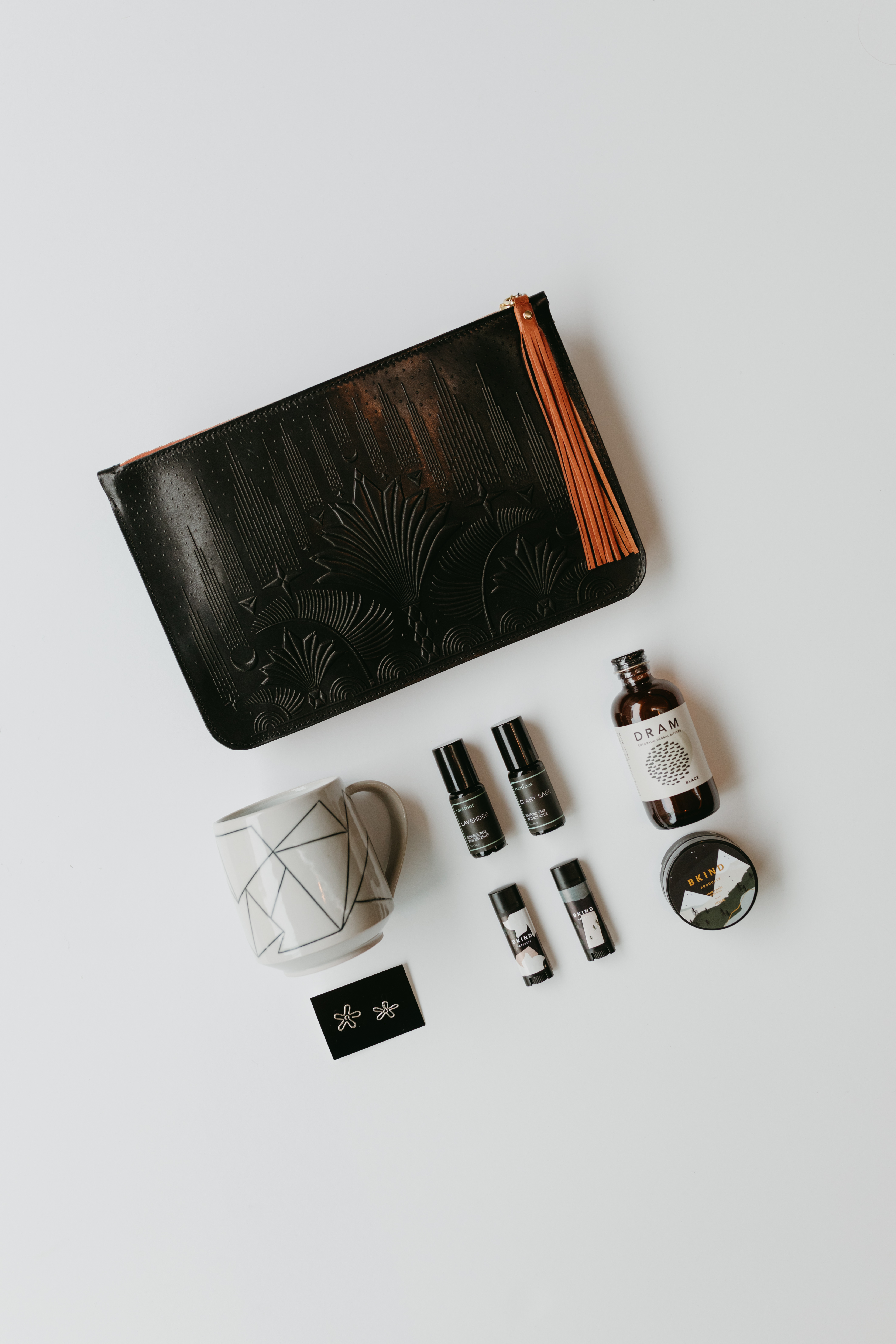 A black leather pouch. A white mug with geometrical patters. A pair of silver floral wire earrings. A bottle of bitters from DRAM. Hand balm and chapstick from B KIND. Fragrance roller from rootfoot in lavender and clary sage. 