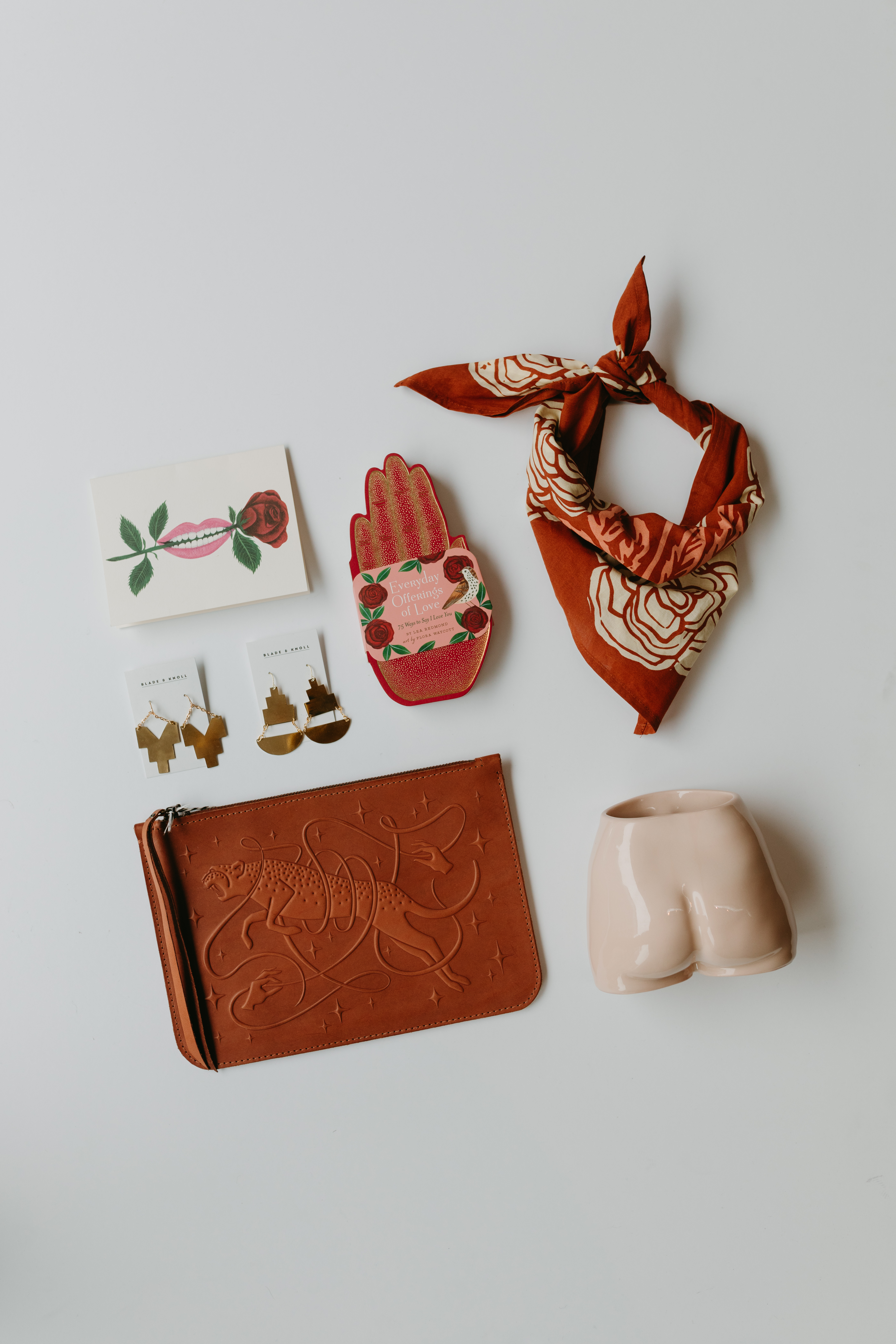 Seven objects on a blank white surface. A red ascot. A succulent potter in the shape of a persons buttocks. A leather pouch with an imprinted leopard. Two golden earrings. A postcard depicting a mouth biting a rose. A red hand with the text “Everyday Offerings of Love.”