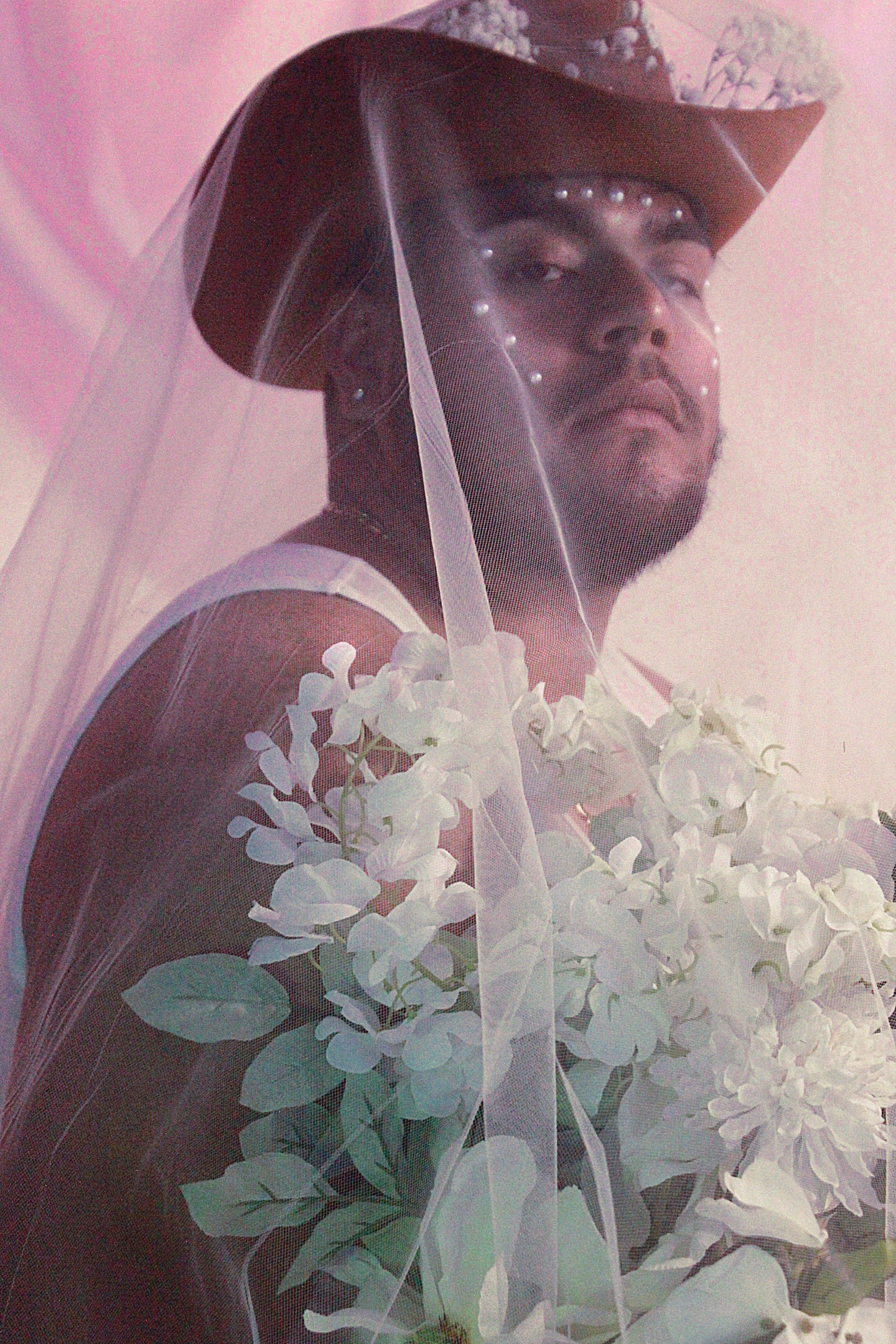 A portrait of Ulises, wearing a white tank top and a red cowboy hat, surrounded by white flowers and covered by a soft pink veil. The look on Ulises face is soft yet strong