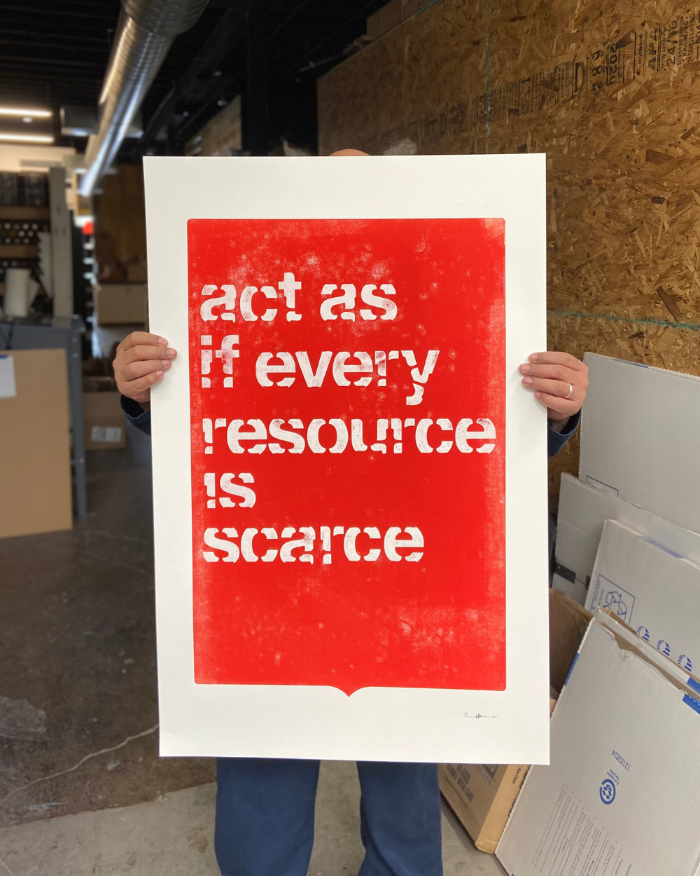 Photo courtesy of Rick Griffith. [Image description: a person holding a large print that covers their face, torso, and thighs. The poster is red with a white border, there’s white text on it that reads “act as if every resource is scarce” in all lower case letters. Pieces of the lettering look cut off. The image is captured in what looks like a warehouse or studio.]