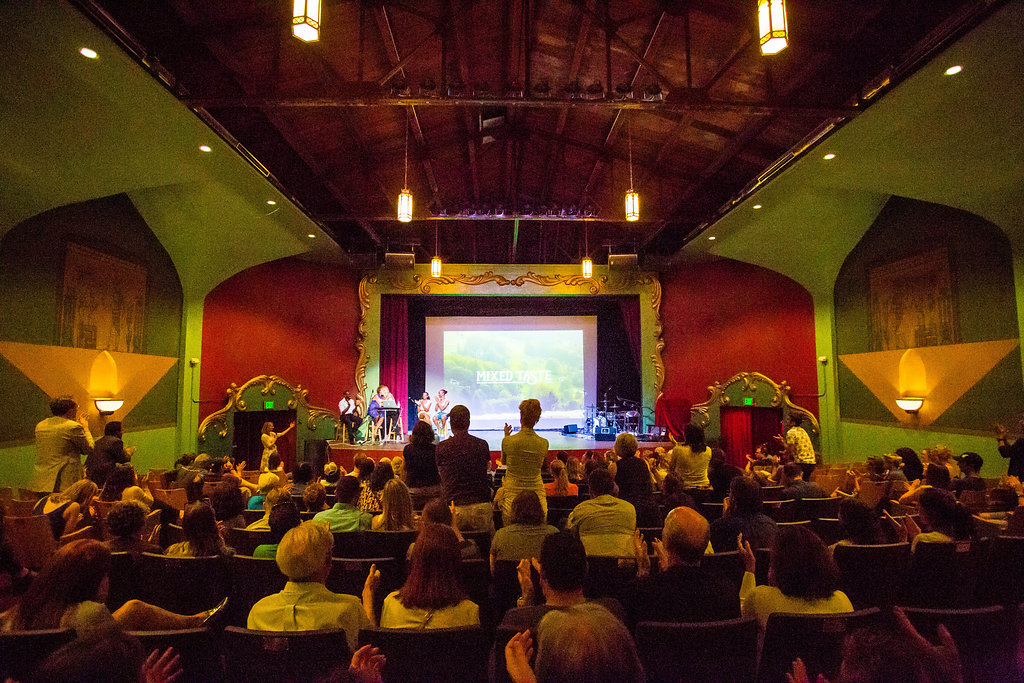 Back of a crowded performance space. Members in the audience are facing the stage: some are sitting, some are sitting and clapping, others are standing and clapping. There’s a large screen tucked between red floor length curtains and displays a presentation that has text that faintly reads, “MIXED TATSE”  