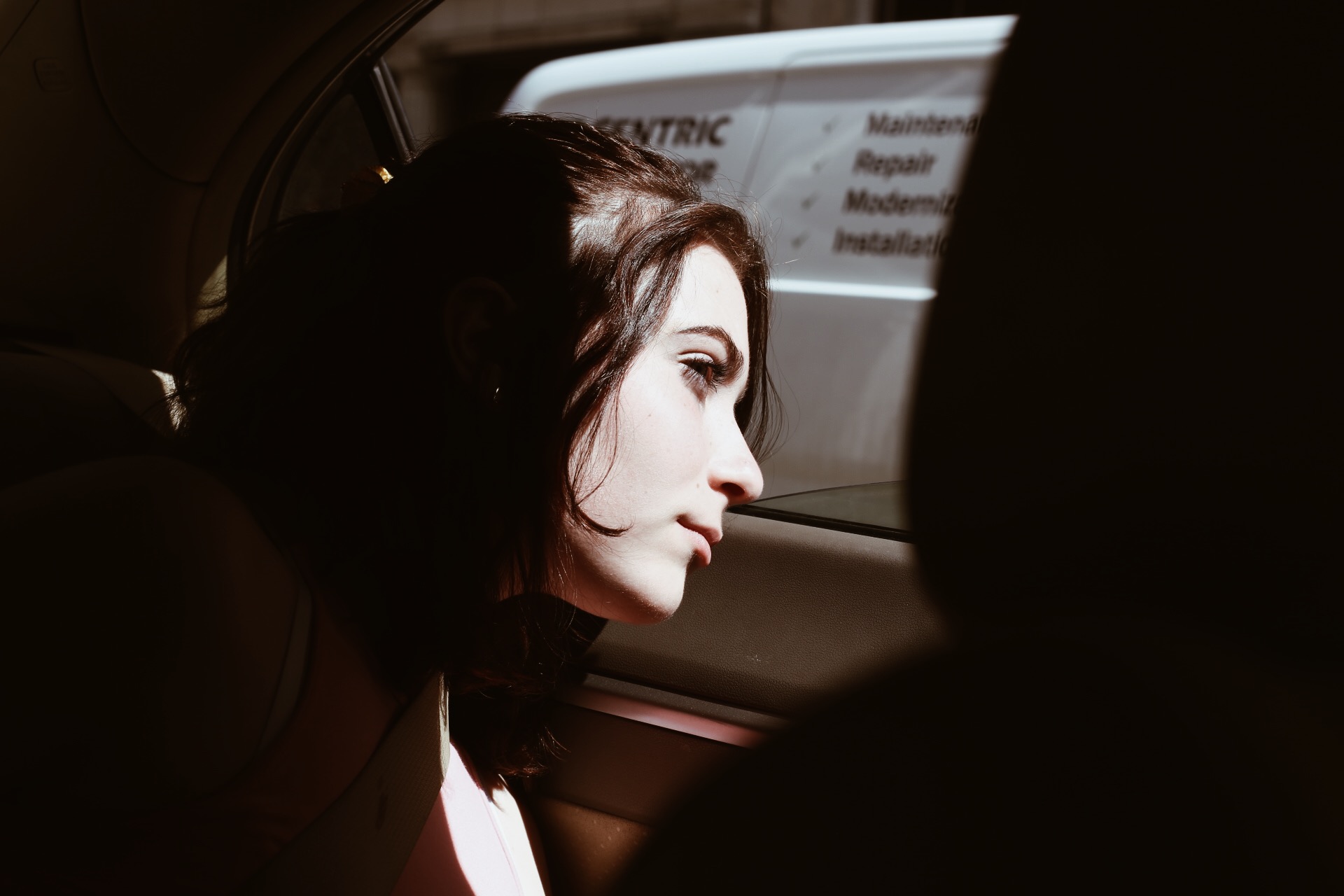 A young woman in a car leans her head to look out a car window. Only her face is lit. 