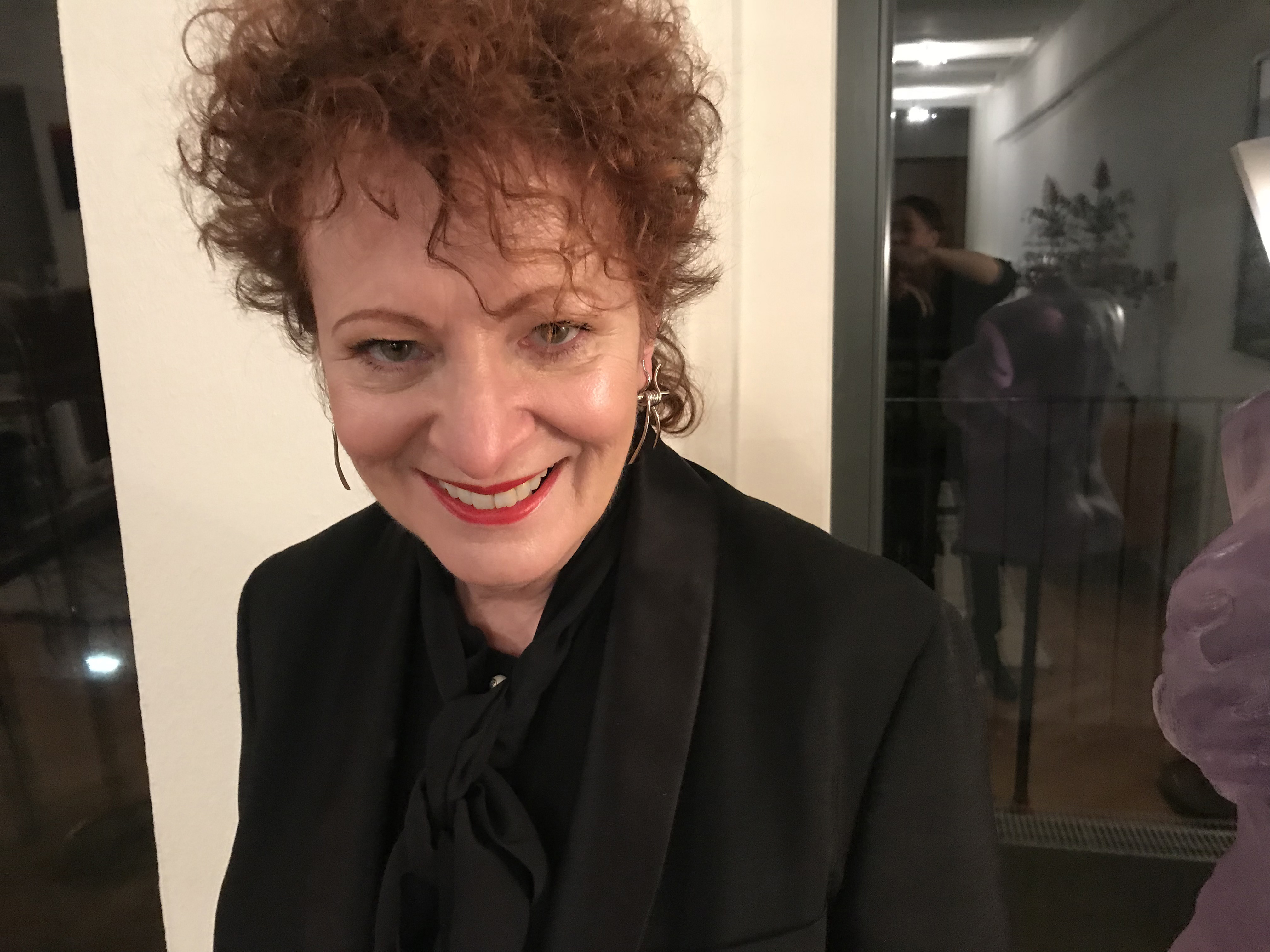  Portrait of Nan Goldin in an unidentified indoor space. She is wearing a black blazer with a black ascot. Her curly auburn hair falls across her forehead as she smiles warmly into the camera. Behind her is a window in which sculpture of a woman’s torso is visible. 