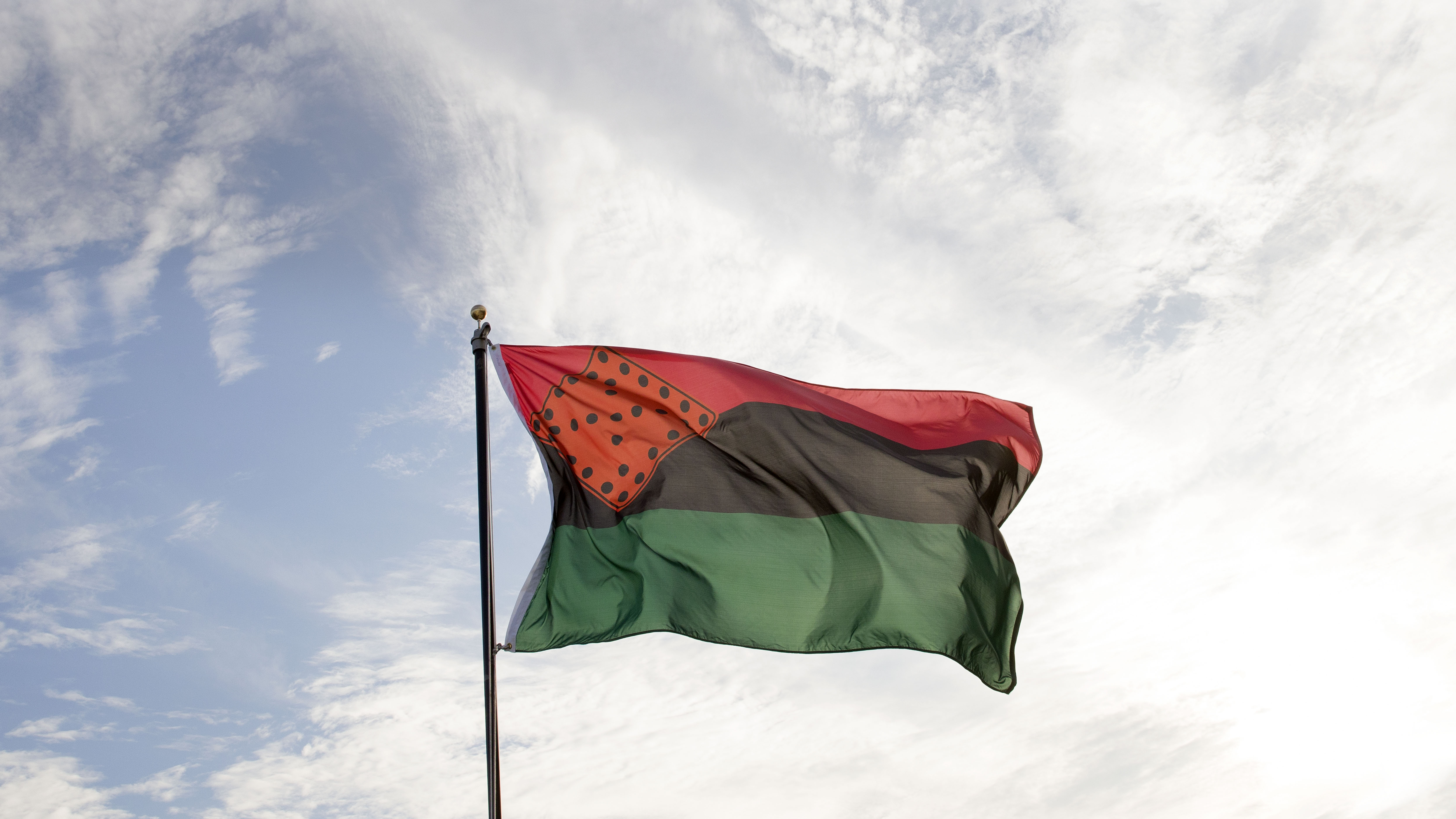 Nari Ward, Breathing Flag, 2017. Nylon flag. A Pan-African flag flies against the sky. It is equally split in red, black, and green colors horizontally. An orange diamond with black dots rests in the upper left corner. 