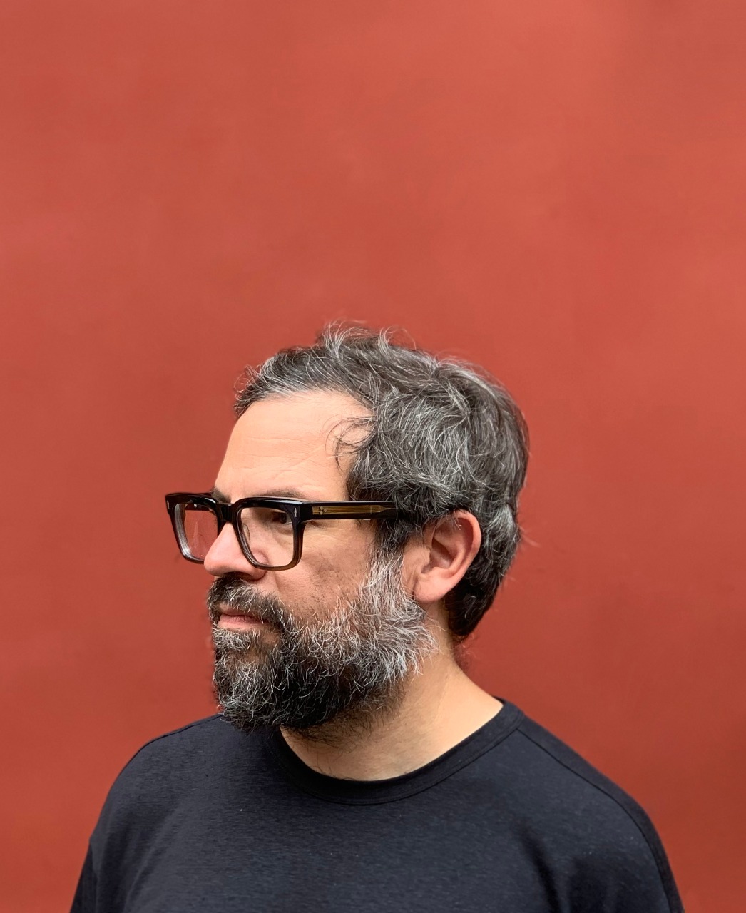 A portrait of Pedro Reyes against a bright red background. He is wearing a black t-shirt and glasses with thick, black frames, and his graying hair rests in a tousled fashion atop his head. 