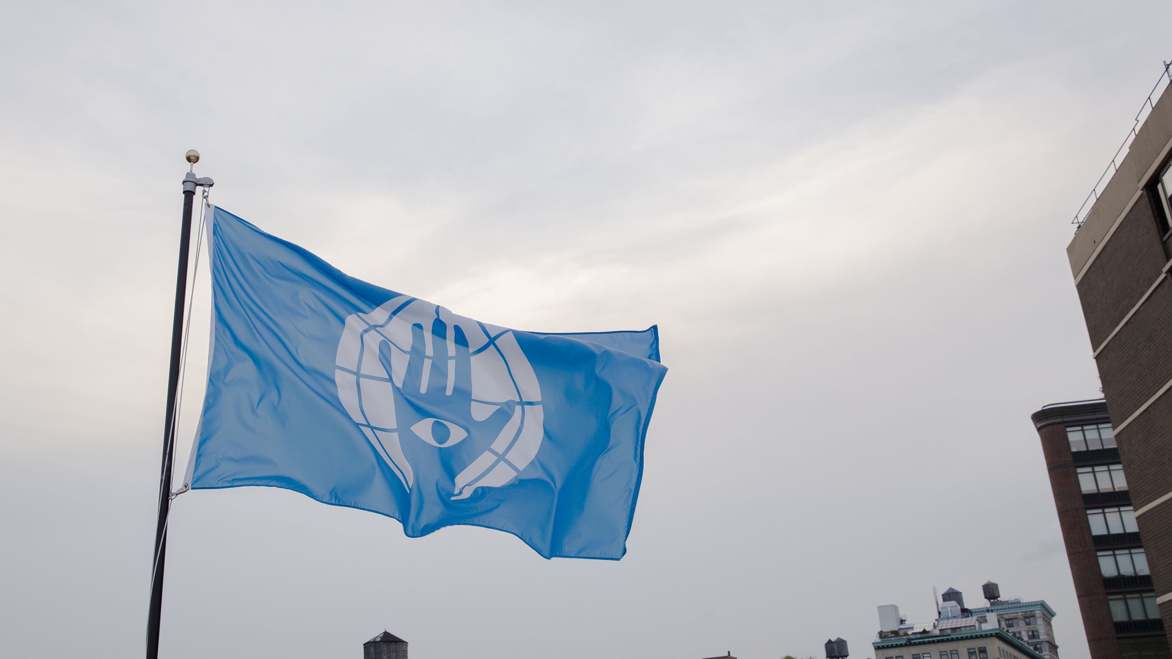 A cerulean colored nylon flag flying in a light blue, almost white sky. The flag has a palm on it, which has an eye on the center of the palm. Around the hand is a circular design. 