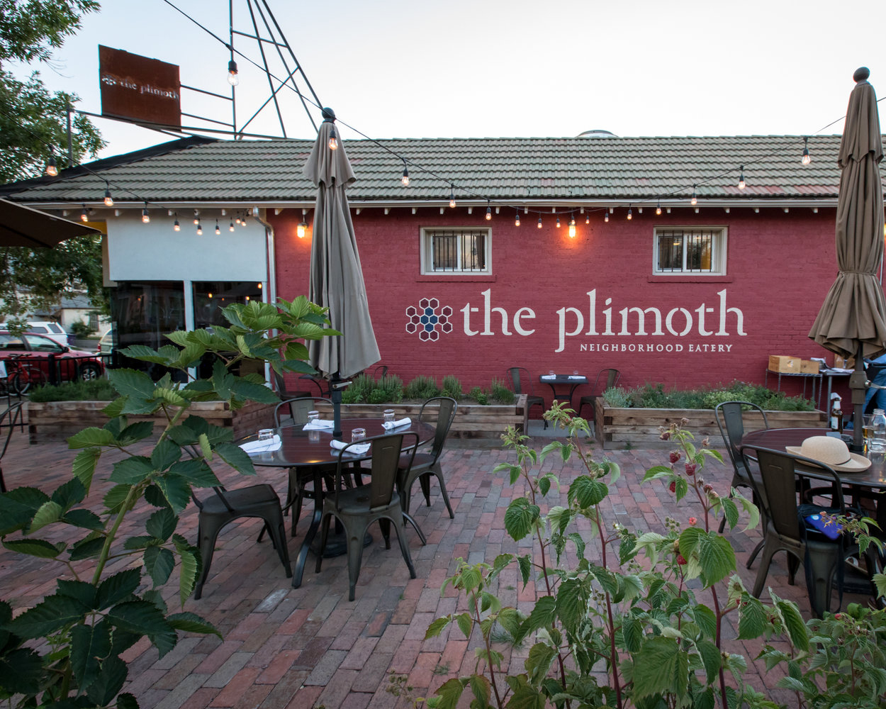 A red building with the words The Plimoth Neighborhood Eatery during the day. There are metal tables outside on red brick below string lights. 