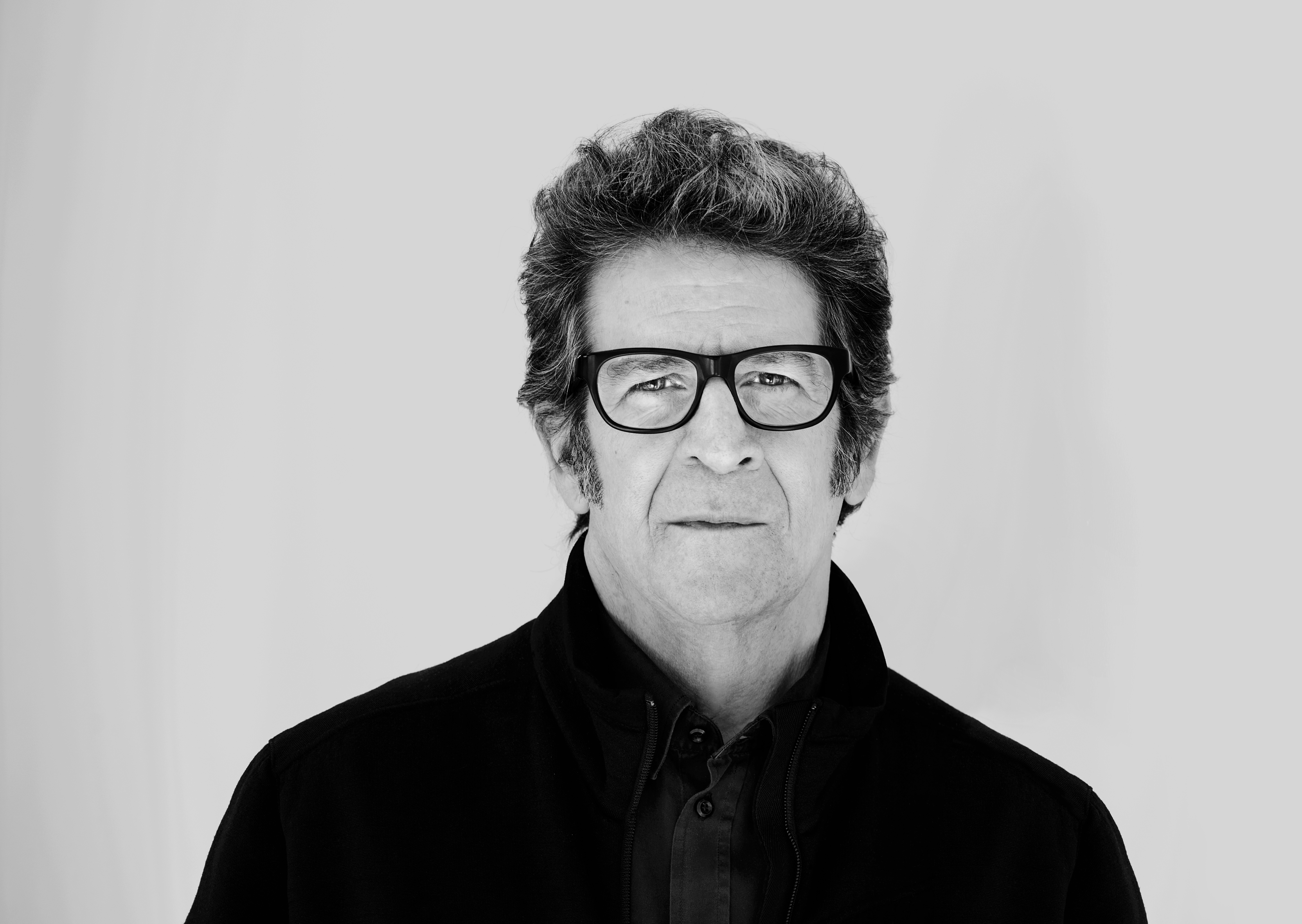 Black and white portrait of Robert standing against a plain wall. He has a serious look on his face, is wearing glasses and  a jacket over a button up shirt. 