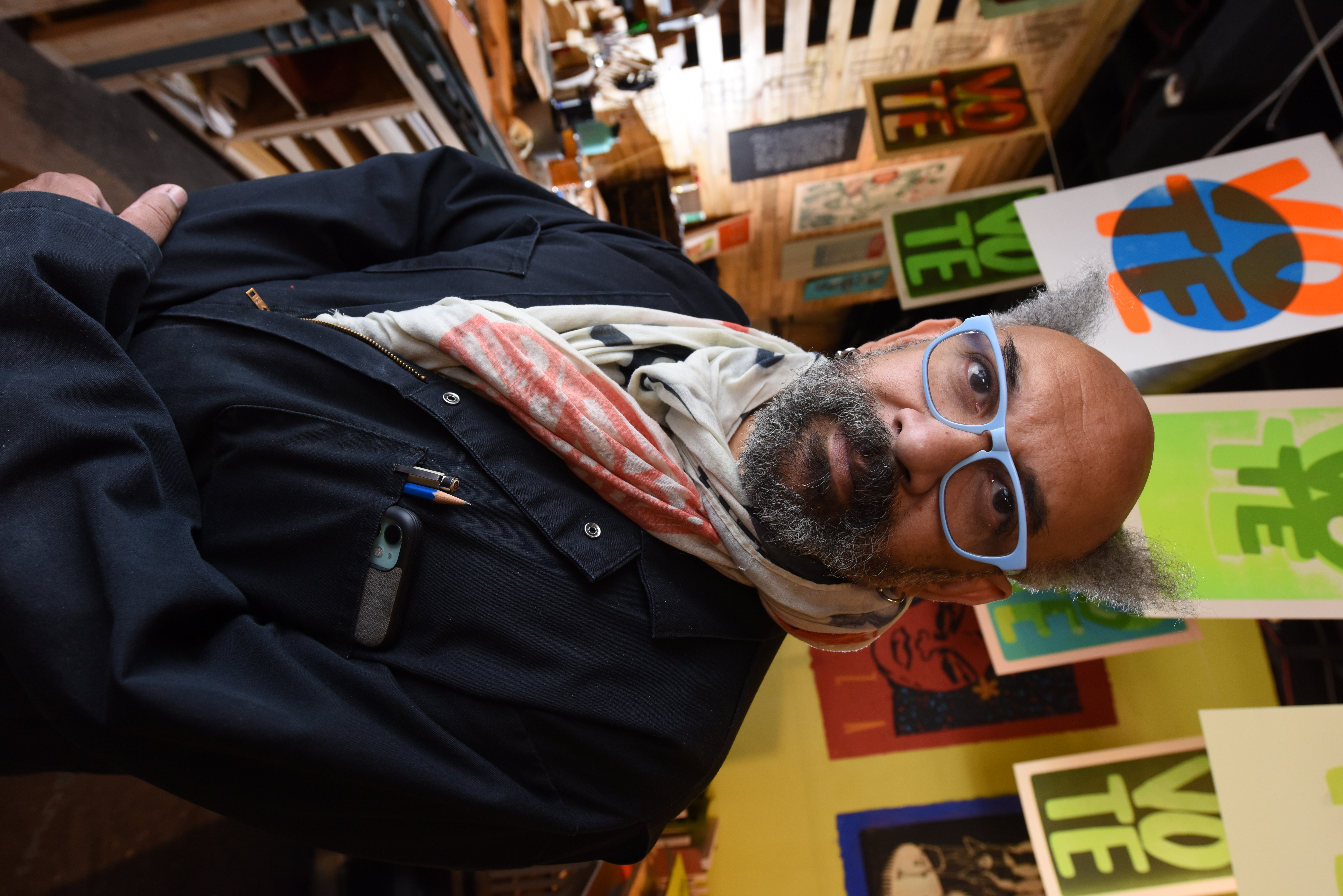 Image of artist Rick Griffith. He wears periwinkle glasses, a scarf, and has a beard. He stands in front of colorful posters that say "Vote" on them.