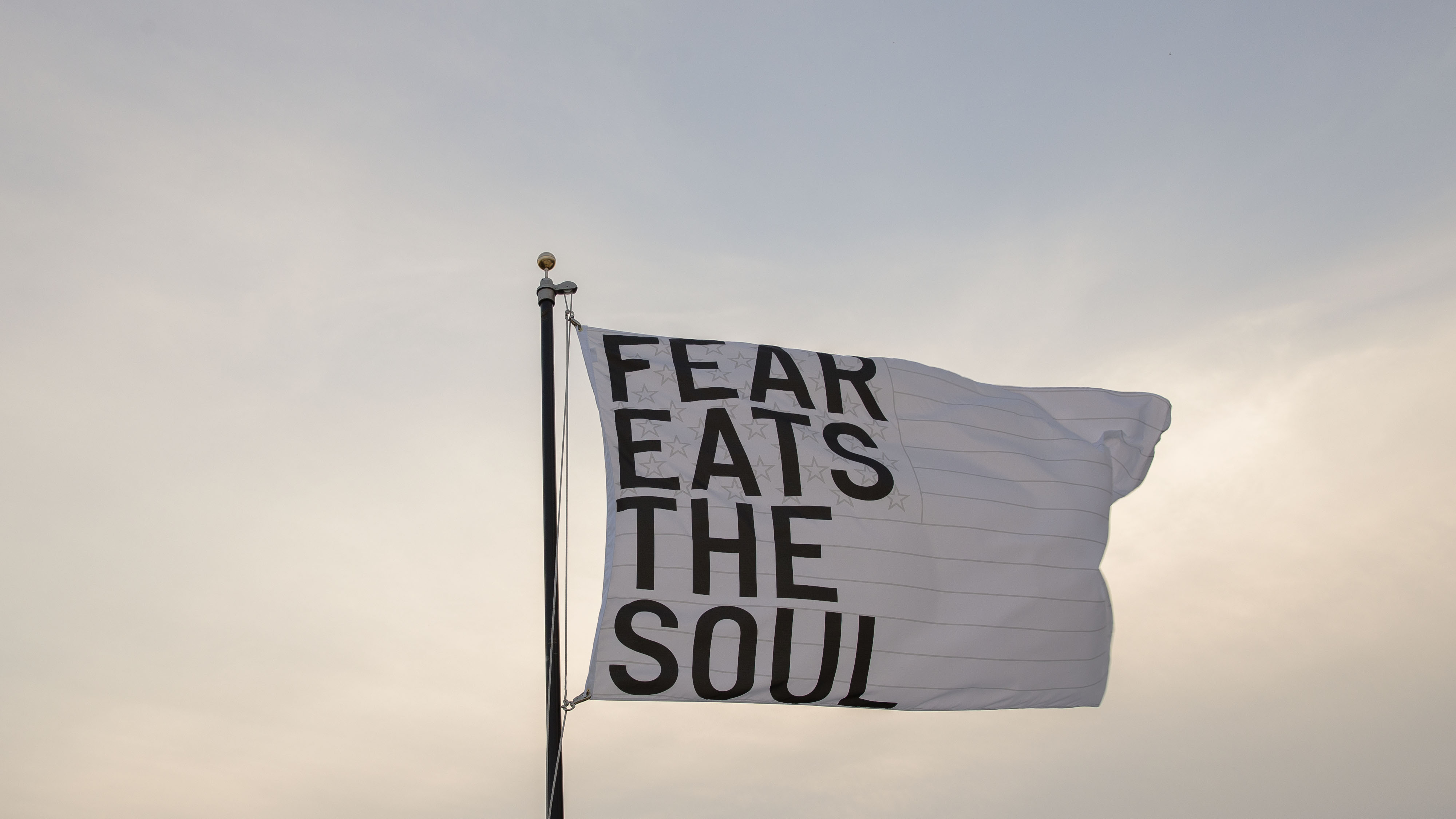 A white flag flying the sky that reads, “FEAR EATS THE SOUL” in black, capital letters. 