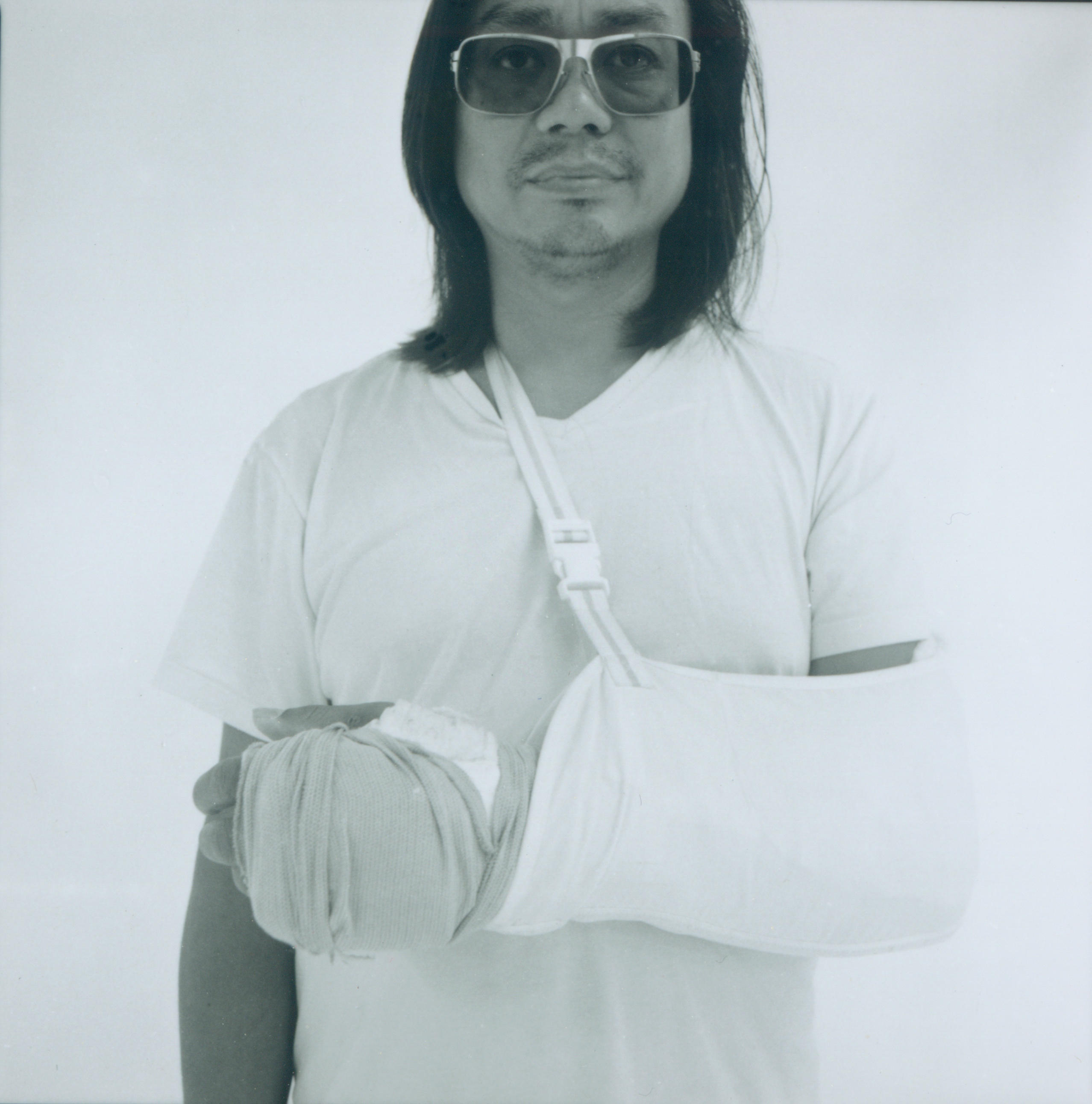 A portrait of Rirkrit Tiravanija, bathed in a light blue hue. Tiravanija is wearing sunglasses, a plain t-shirt, and is looking and facing directly at the camera. His arm in in a sling. 