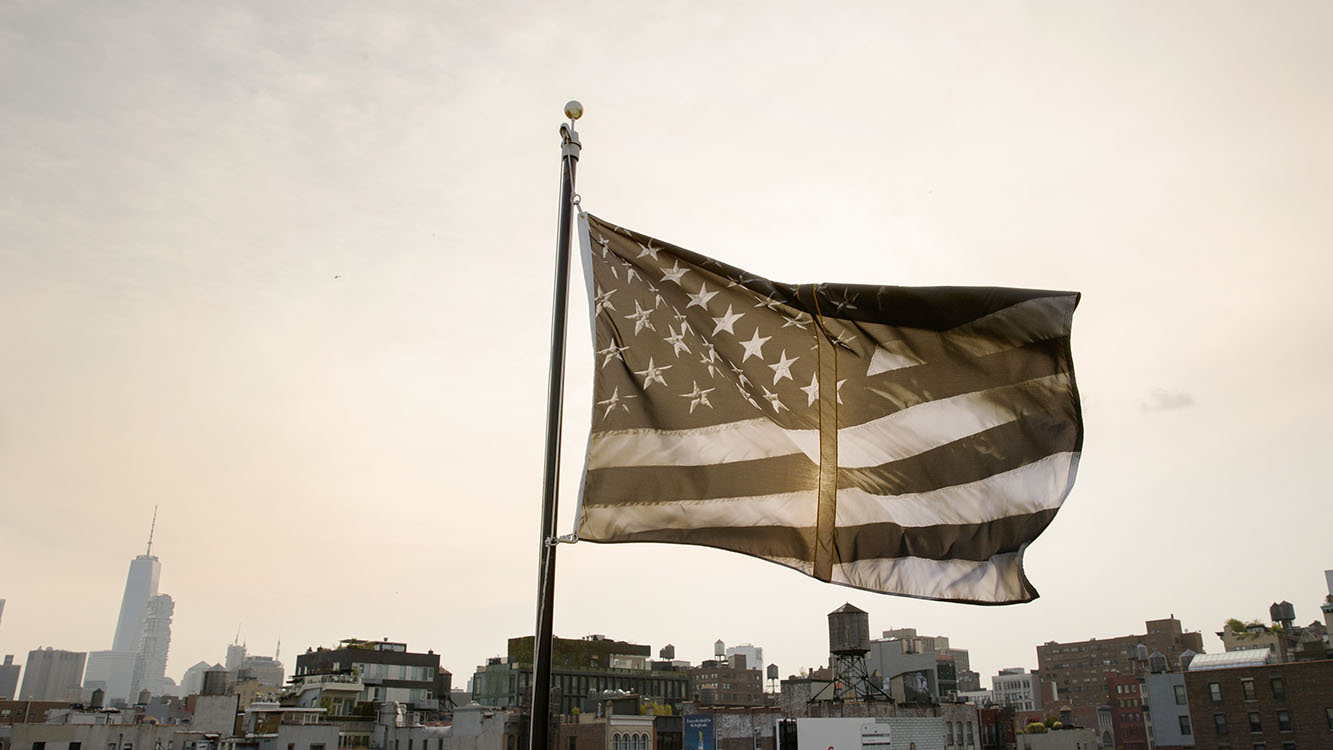 An altered version of the American flag flying in the air. The colors of the flag are muted and there is a thick vertical line in the center on the flag. 