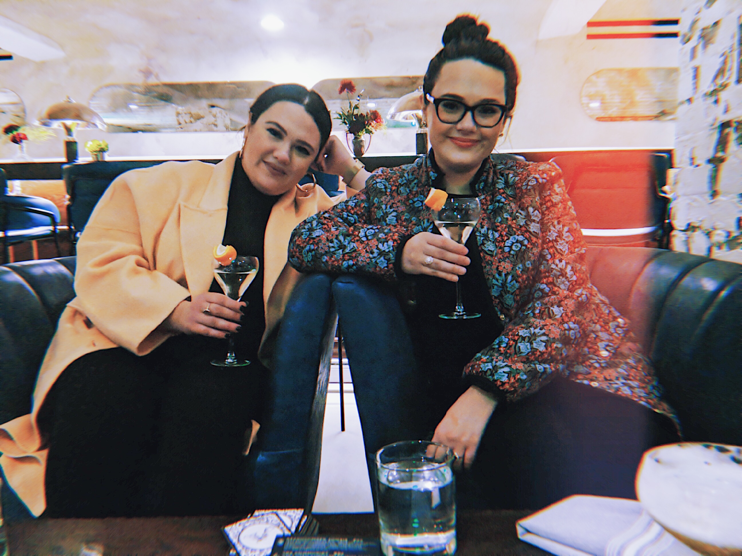A pair of young adult female twins, one more evil than the other, pose for a photo holding two cocktails in a bar. One is in a beige coat with a black turtleneck. The other smiles with malicious intent sporting black framed glasses, and a floral jacket. 