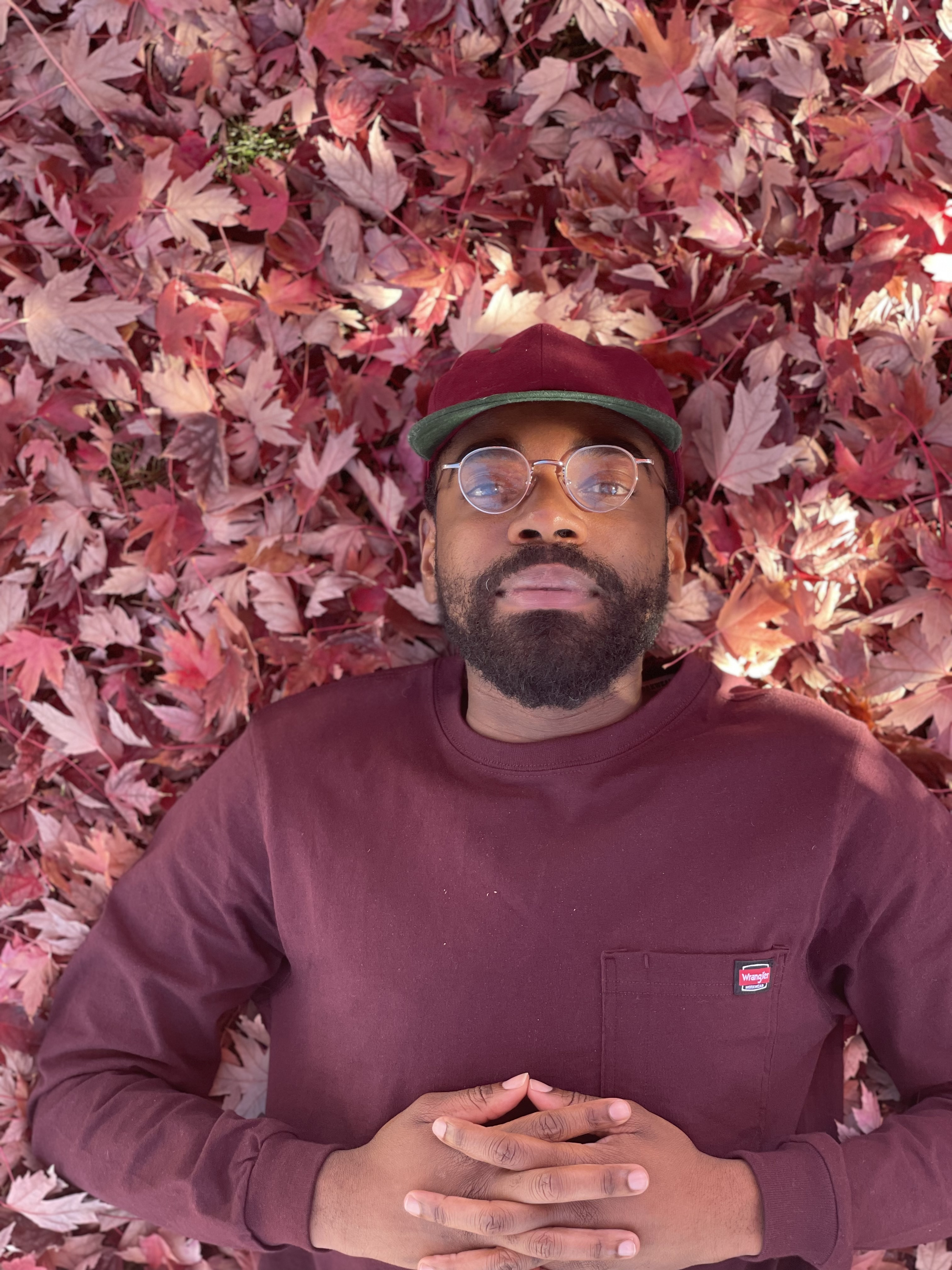 Hip Hop Artist, Schama Noel headshot. Wearing a maroon sweater laying in fall colored leaves