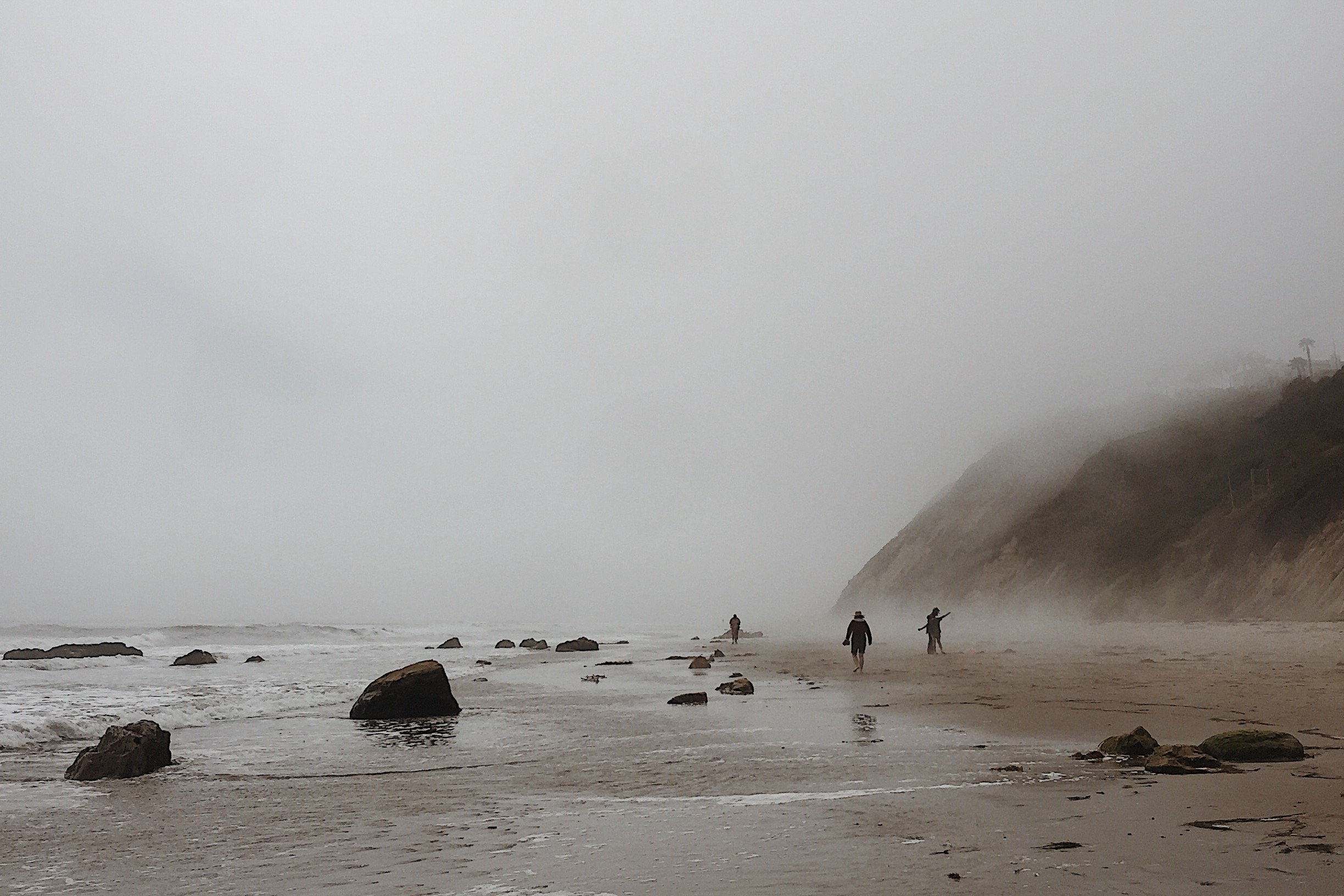 A foggy beach in sepia. There is a cliffside in the distance and four people walking the shore.