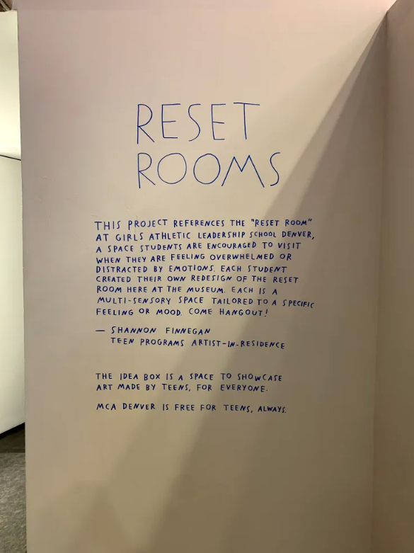 Blue text is written on the wall. It reads: RESET ROOMS This project references the Reset Room at girls athletic leadership school Denver, a space students are encouraged to visit when they are feeling overwhelmed or distracted by emotions. Each student created their own redesign of the rest room here at the museum. Each is a multi-sensory space tailored to a specific feeling or mood. 