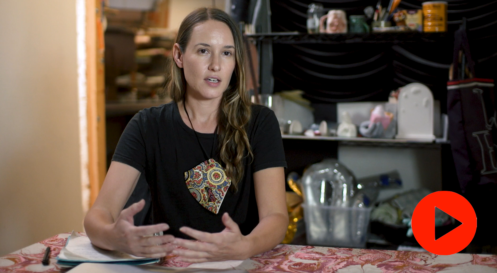 Video still of Laura Shill in a black tee shirt at table. Her brown hair is let down and she is wearing a necklace with a large rhombus shaped pendant painted in an abstract pattern. 