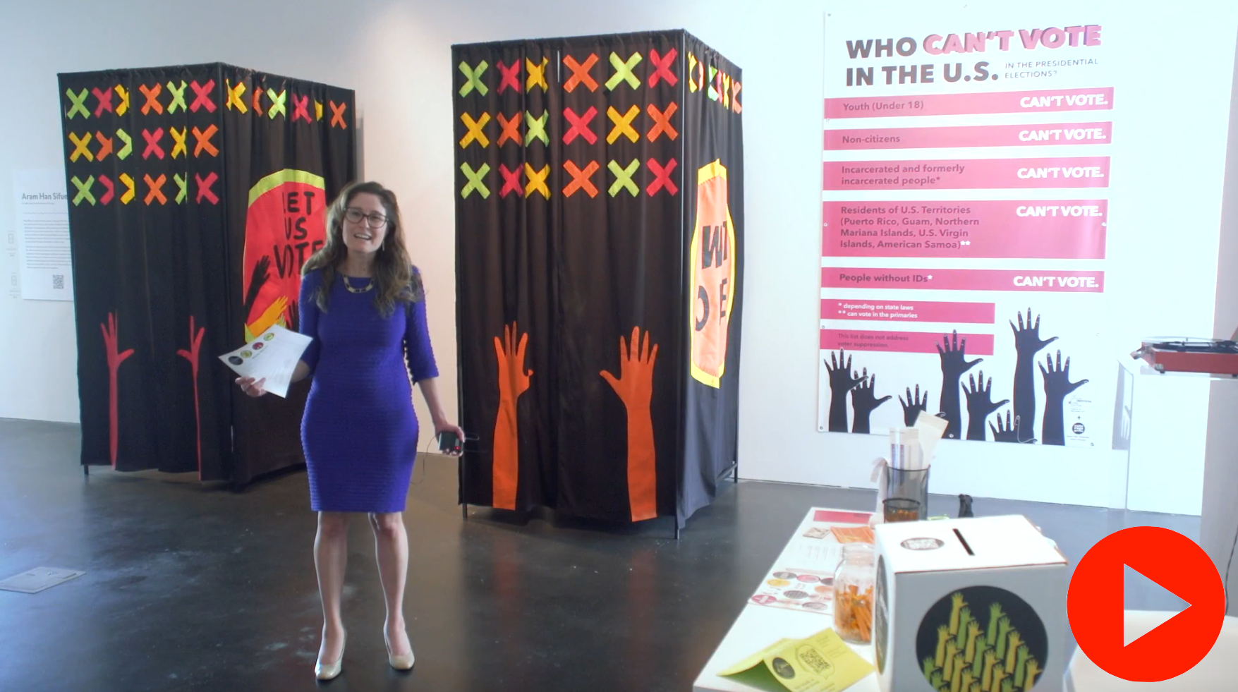 MCA Denver Ellen Bruss Curator, Miranda Lash, stands in Aram Han Sifuentes' installation. Behind her are two voting booths made of black fabric. X’s in red, yellow, and orange cover the fabric. Two hands rise up on the fabric on each booth. On the side “Let us Vote” is in a large circle. 