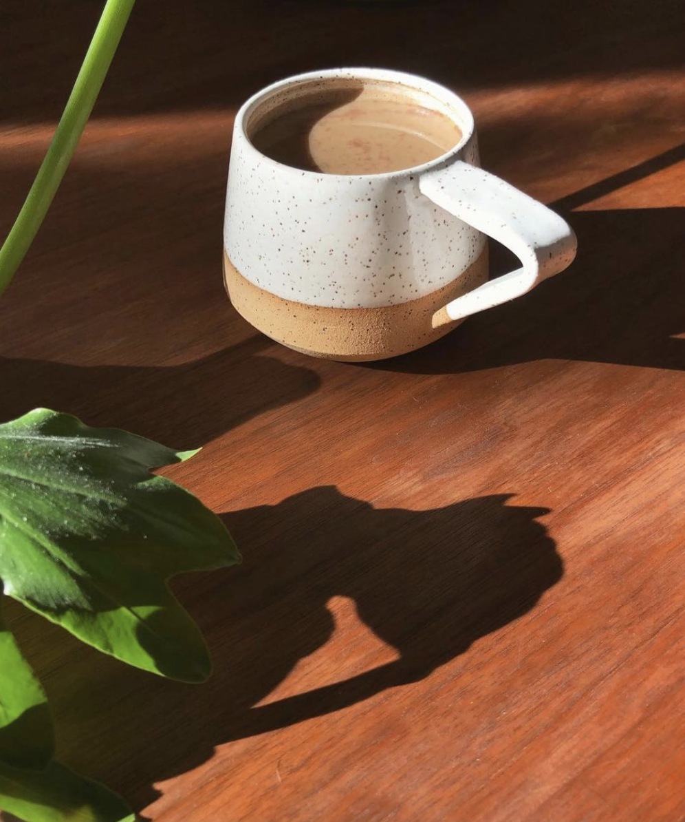 Moody image of a single mug bathing in sunlight on a wooden table. On the left side of the frame is a green plant. The mug is predominantly white with black speckles, with a light brown stroke of color towards the bottom. 