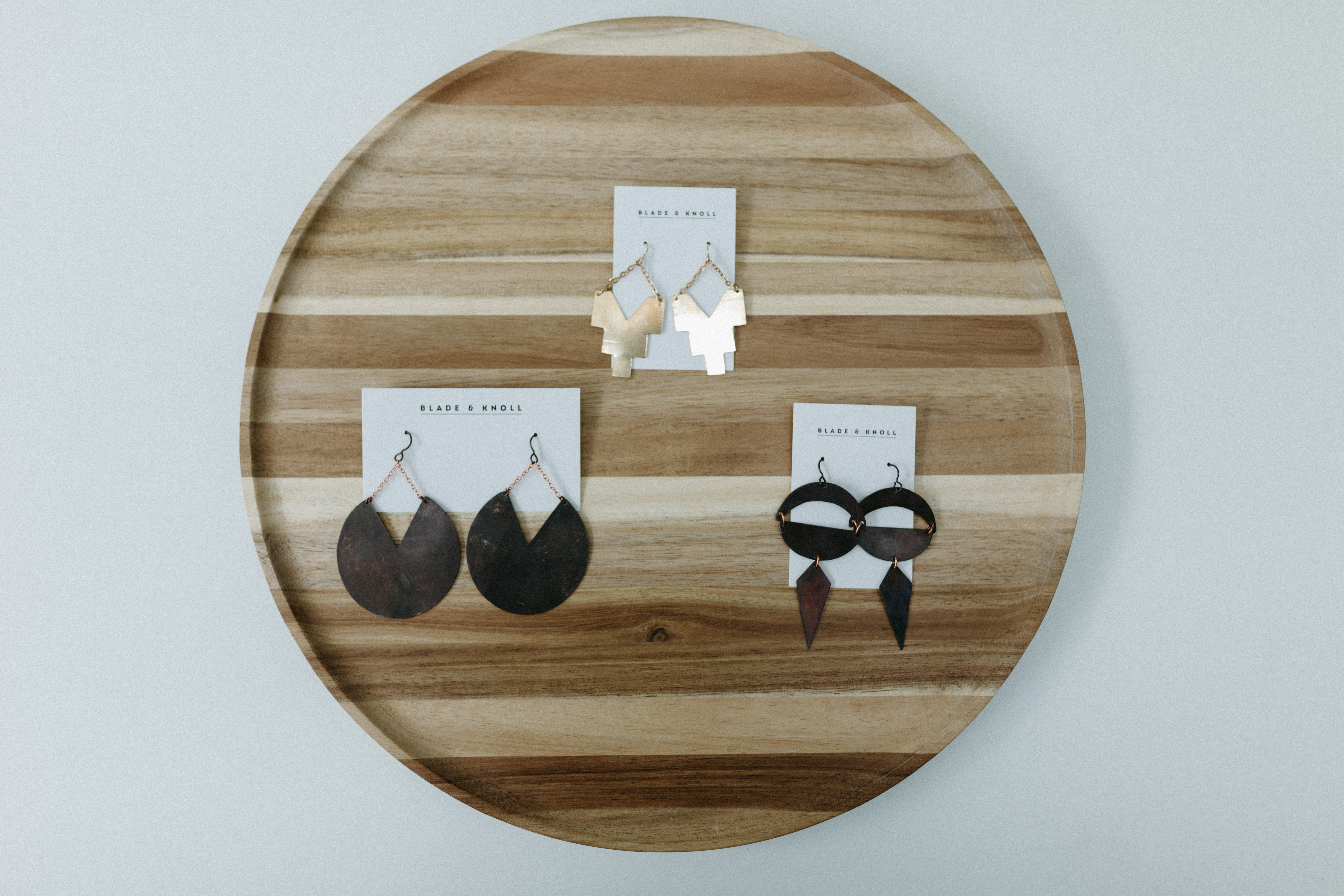 Three sets of earrings from Blade and Knoll rest on a round wooden tray. They are made from metal. Two are dark gray and one is golden. 