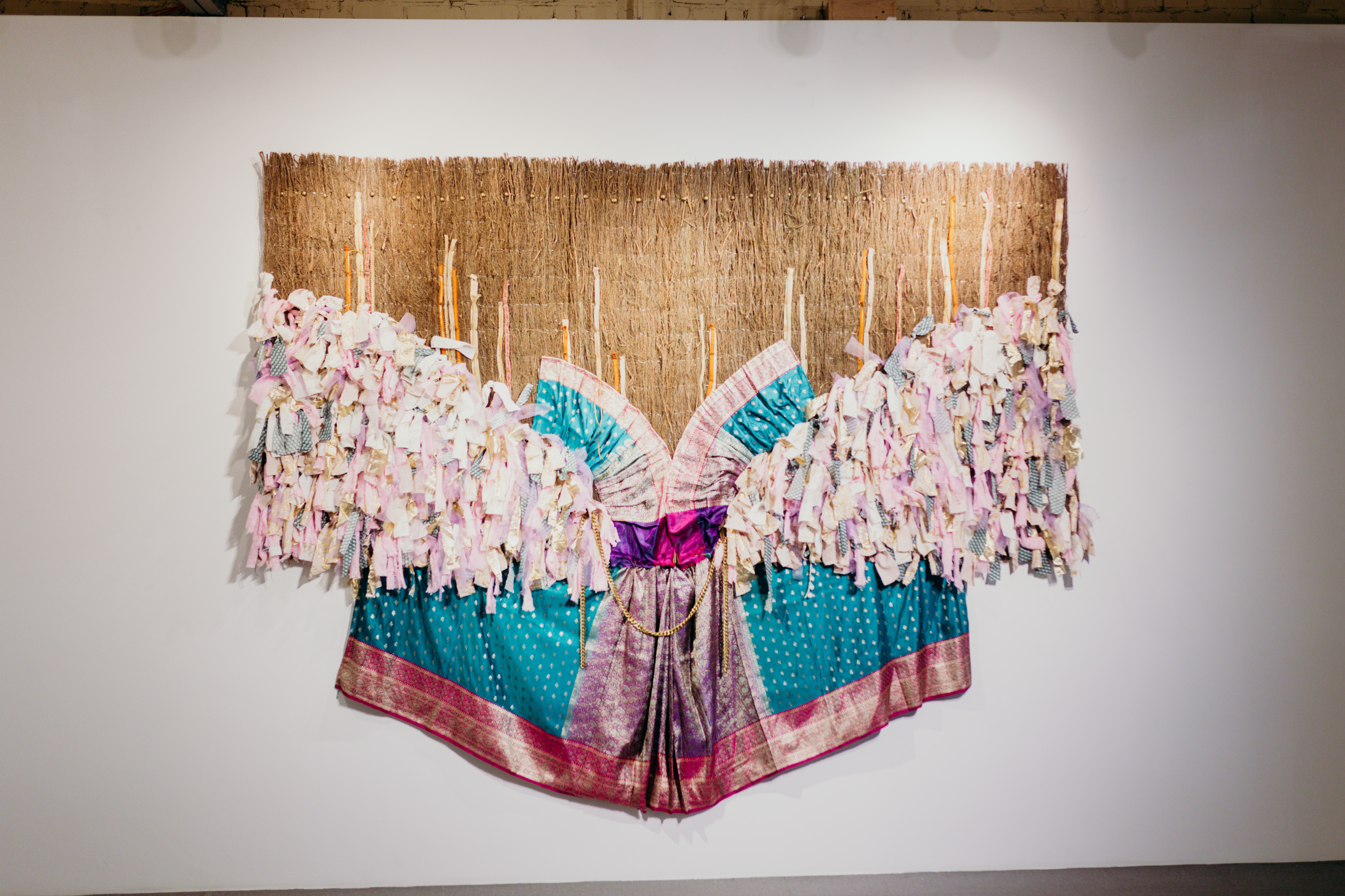 A flat sculpture made from pink and blue frilled fabrics hangs on a white gallery wall
