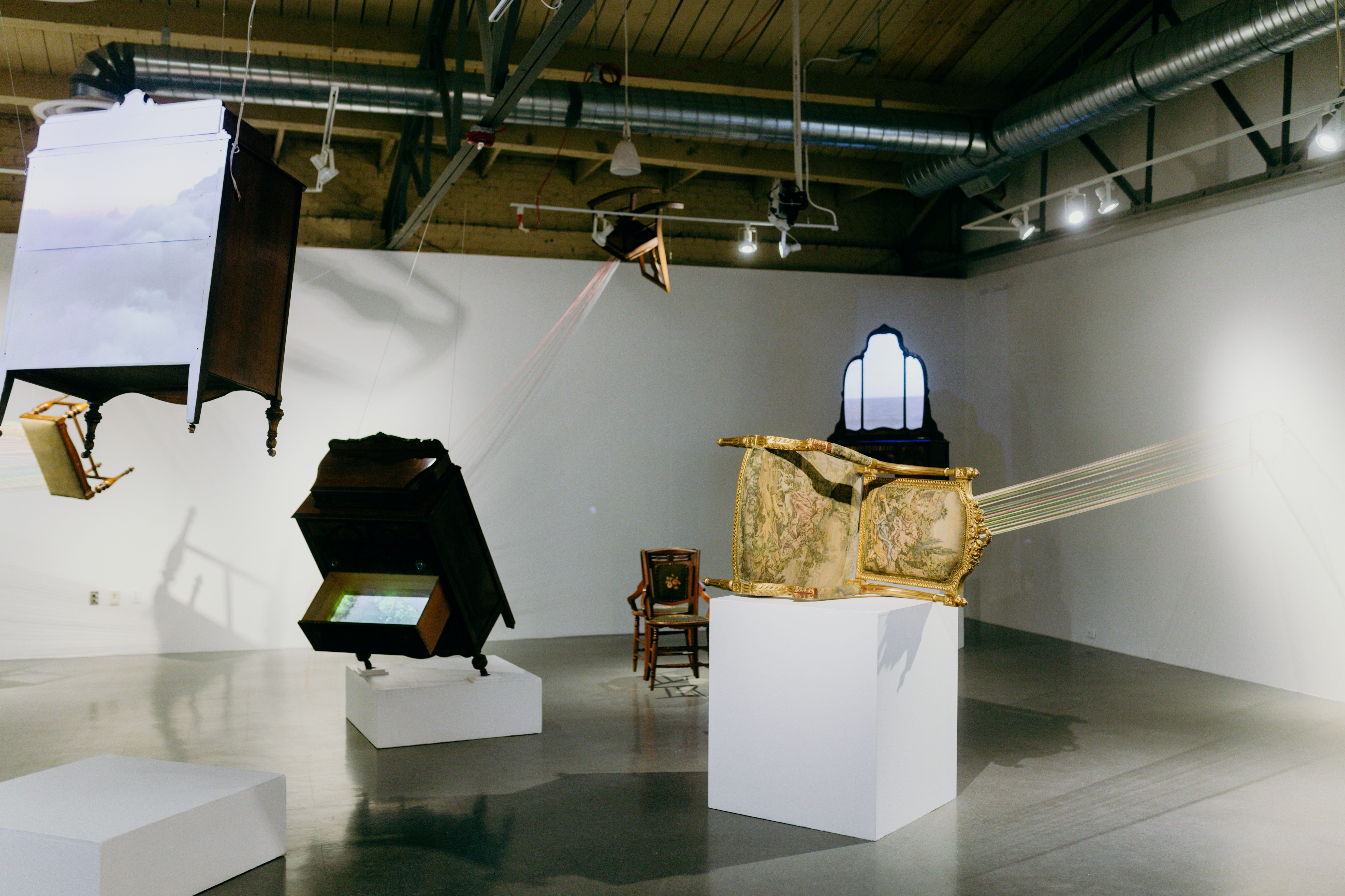 Installation shot of an interior gallery space. There are pieces of furniture suspended mid fall upon white pedastals. ﻿