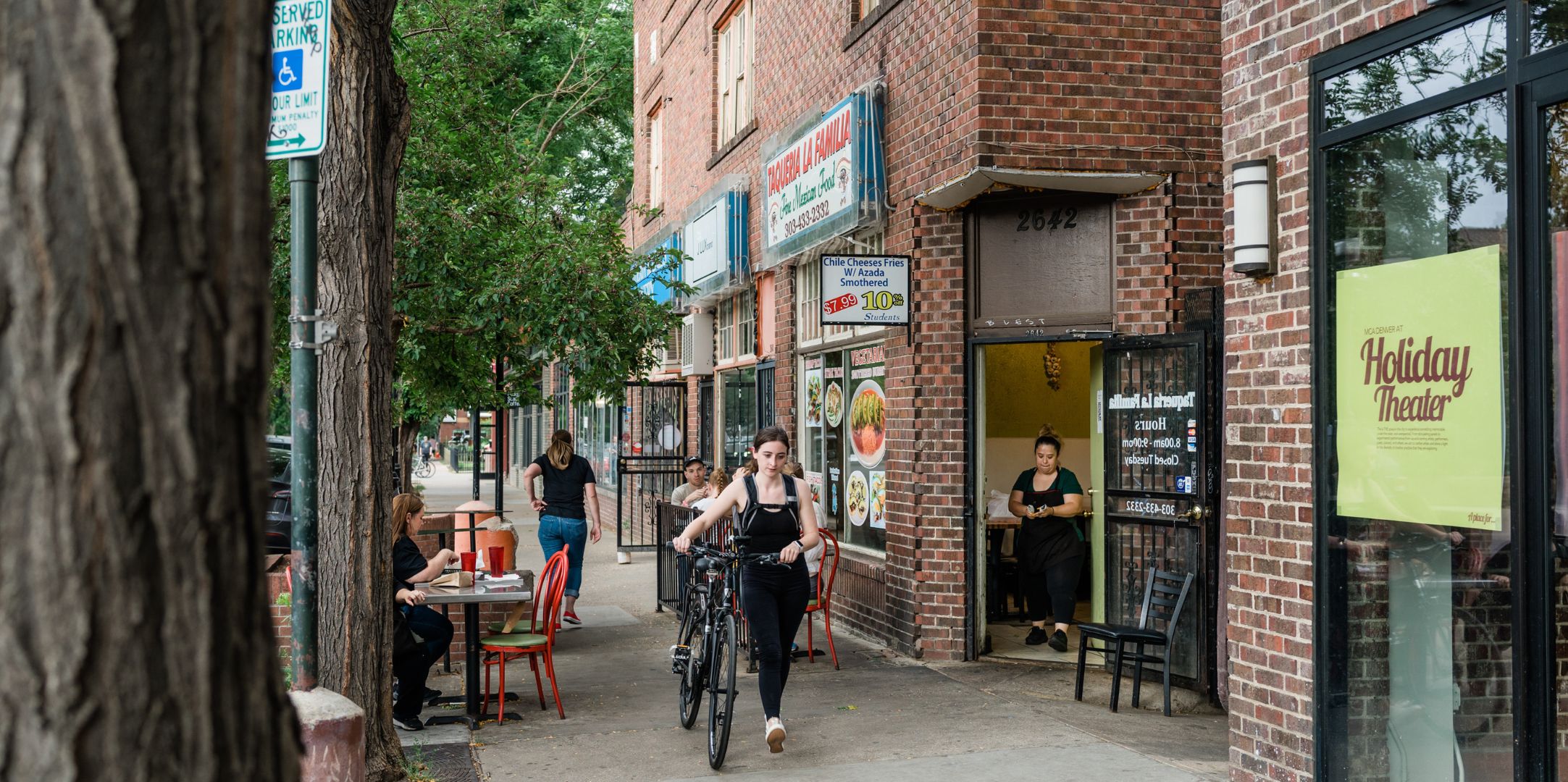 Person walking their bike on a sidewalk near a brick building. There are storefronts lined up along the sidewalk, and people sitting at tables outside one of the restaurants in view.