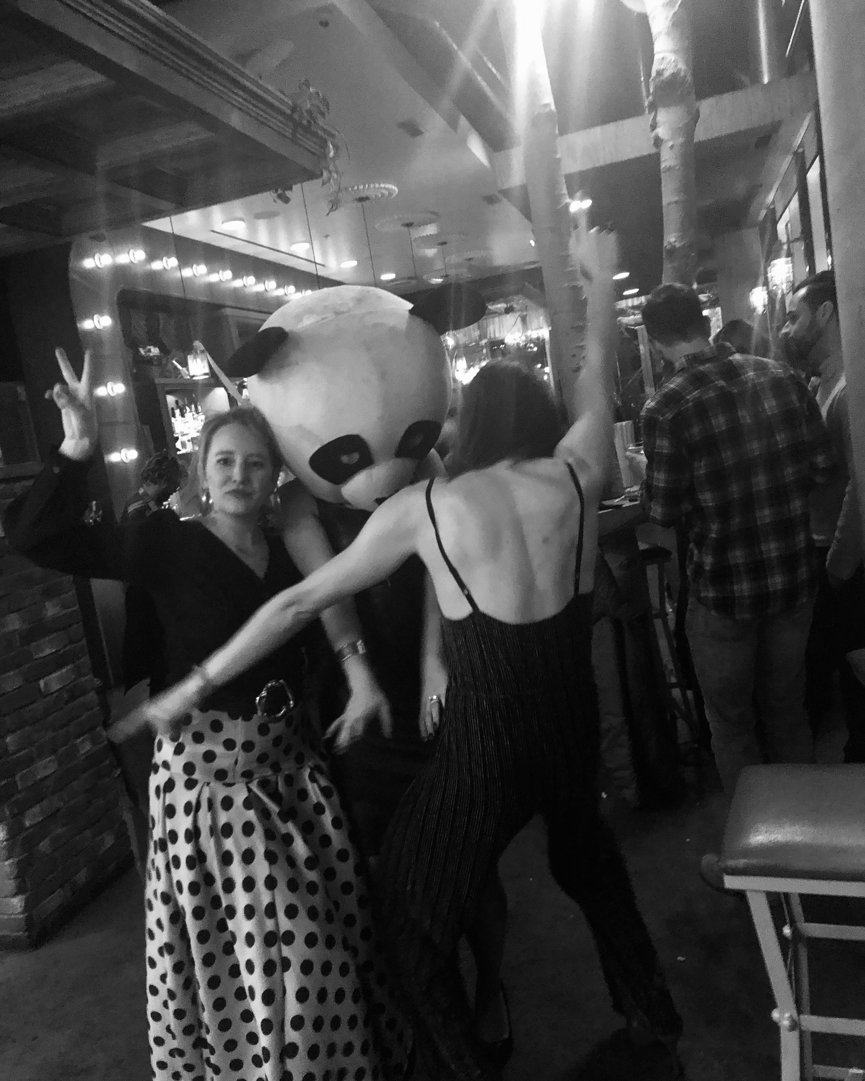 Two young women are in a bar setting posing with a person wearing a panda’s head. One wears a polka dotted dress and holds up the peace sign. The other is mid dance and in a black jump suit. 