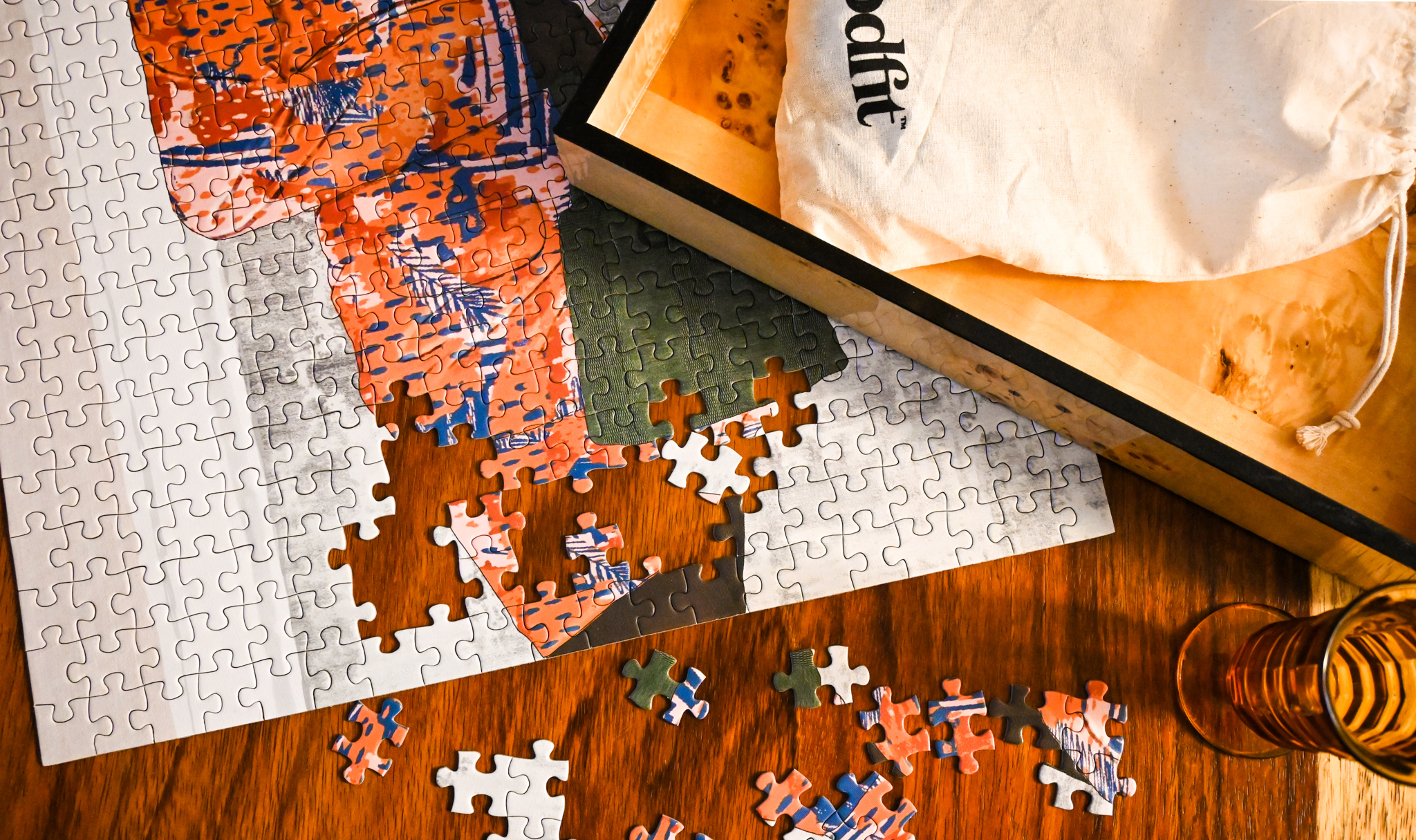 Close up image of a puzzle that is almost complete and is on a wooden table. Many of the puzzle pieces are white, and there is a red and blue printed robe in view. The puzzle’s box and a reusable beige cotton bag lay next to the puzzle. 