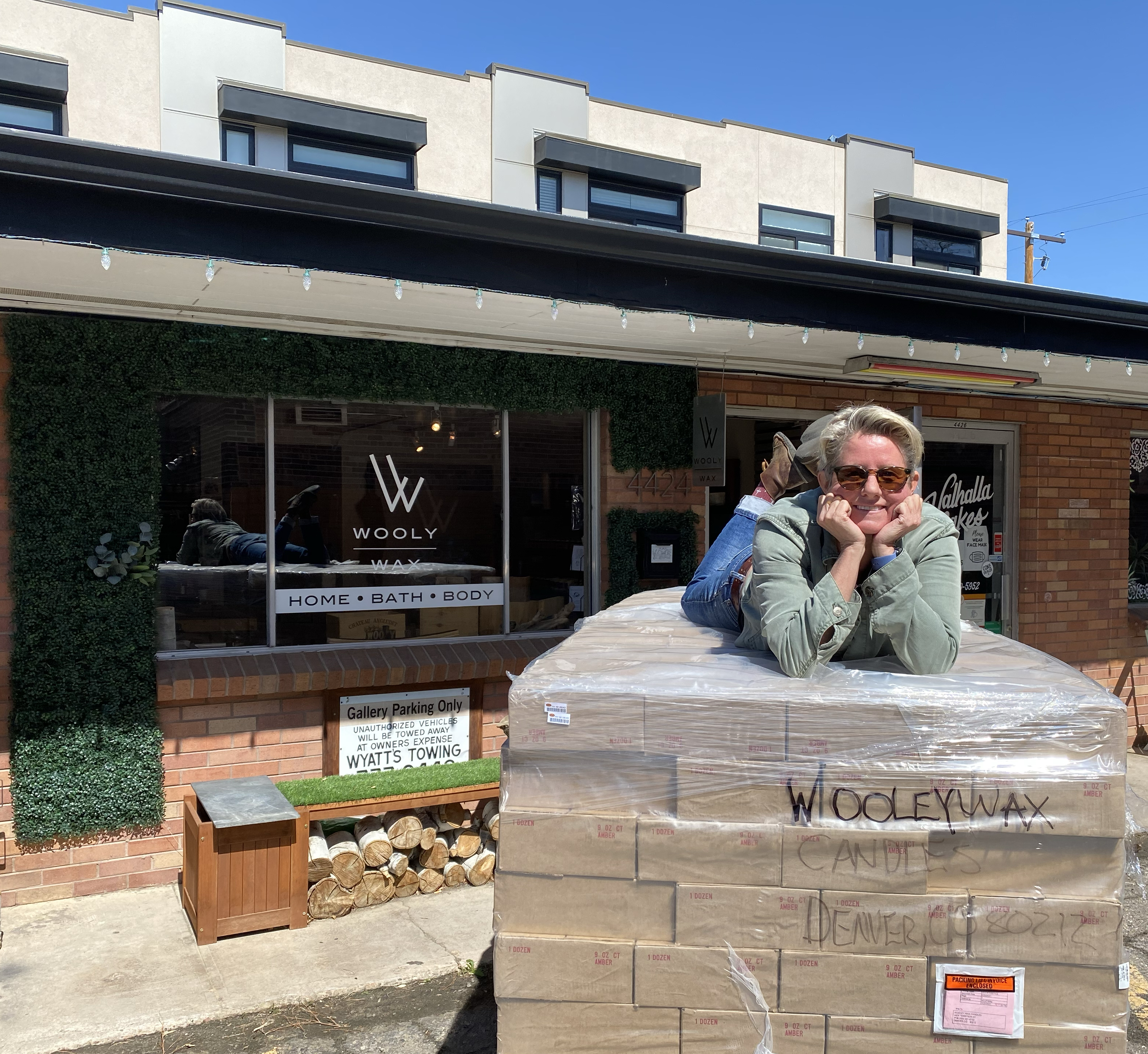 Rachel of Wooly Wax candles laying outside on her stomach on a large and tall stack of boxes outside her storefront. Behind her is a brick building, partially covered in greenery, with the text “Wooly Wax” and “Home, Bath, Body” printed on the window.