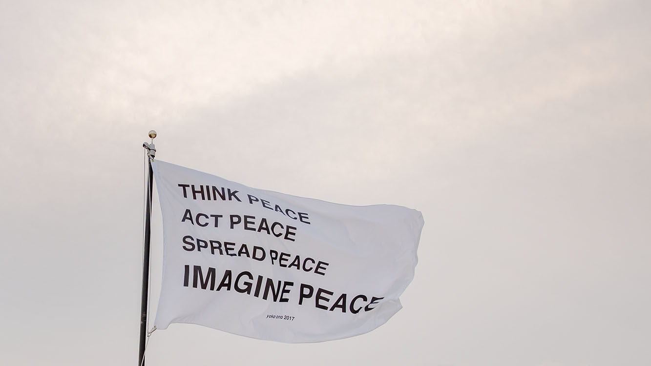 A white flag flying, with the words “think peace, act peace, spread peace, imagine peace” on it. 