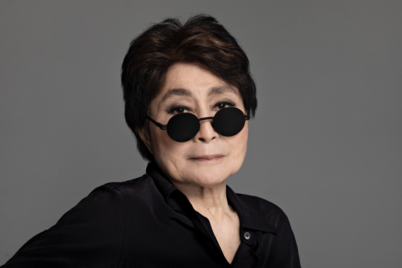 Portrait of Yoko Ono in front of a dark gray backdrop. She has black circular glasses resting on the bridge of her nose. She is wearing a black button up, has on thick eyeliner, and has a small smirk on her face. 