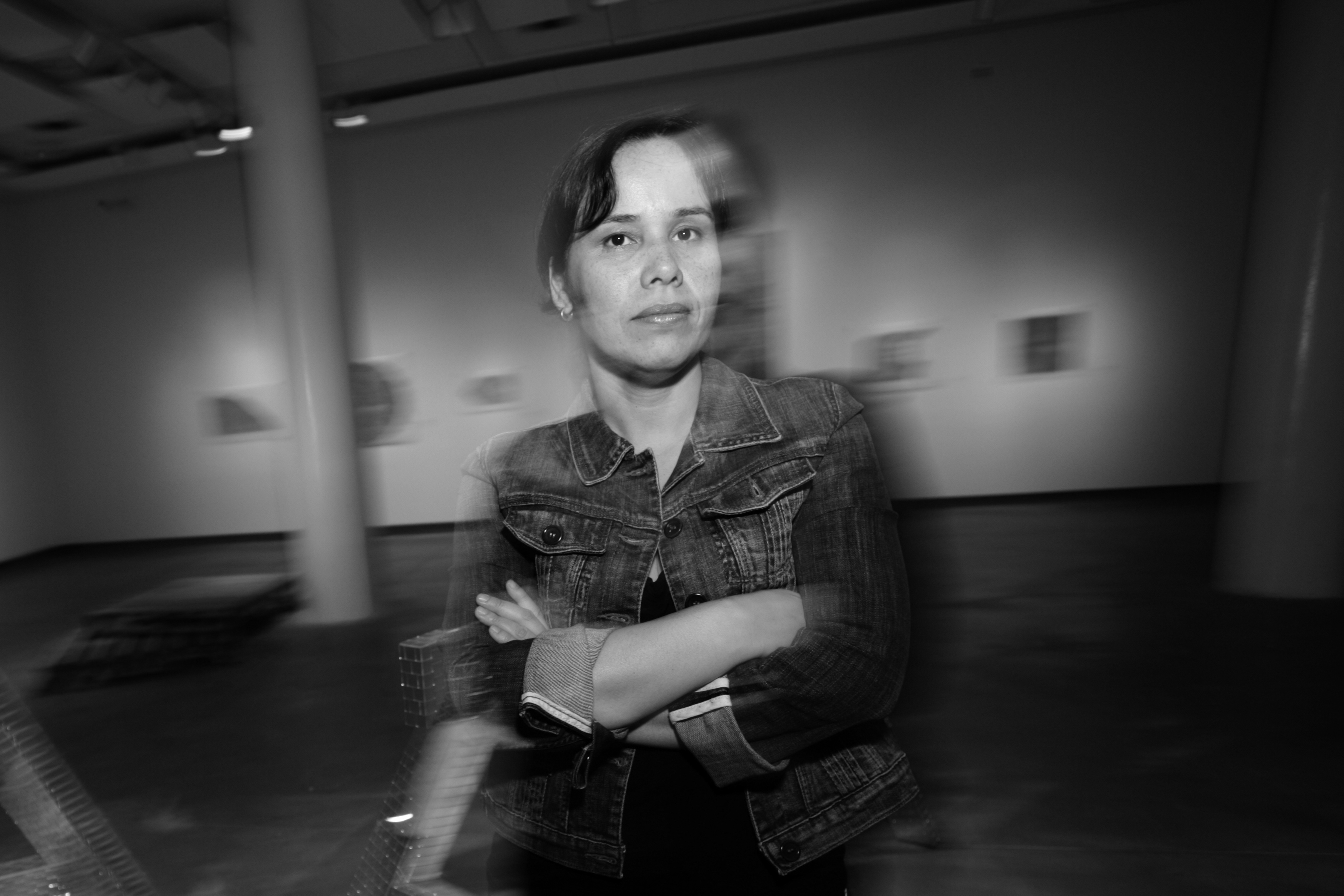 Somewhat blurry black and white portrait of Yumi Janairo Roth. She is wearing a jean jacket, a printed skirt, and has her arms crossed. She has a serious yet soft look on her face and is standing indoors in an unidentified space.