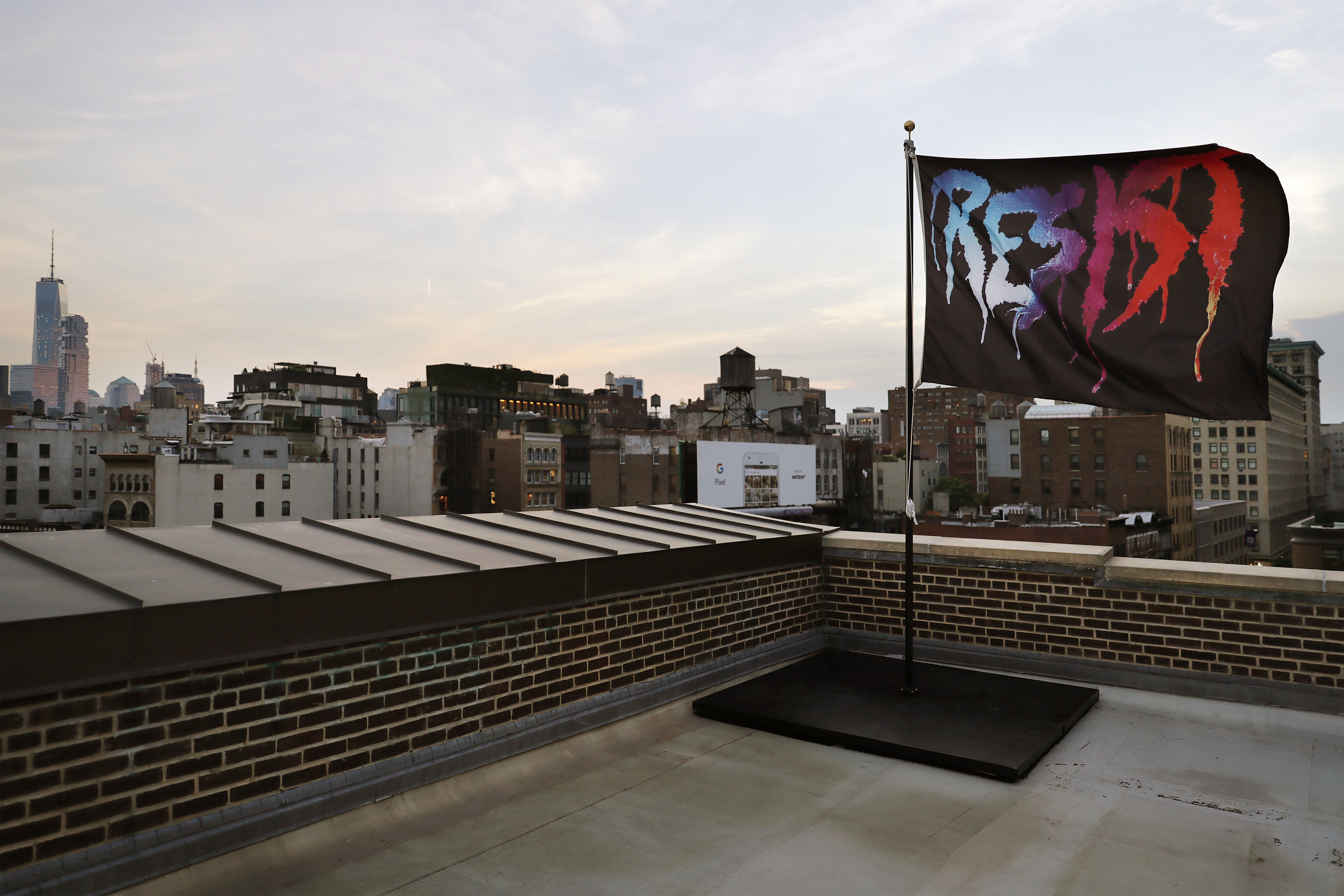 A black flag on a rooftop positioned in the very right of the frame with the word resist in large capital letters on it. The word resist looks almost spray painted, and consists of the colors blue, purple, pink, red, and orange. The flag is flying in the air and surrounded by buildings. 