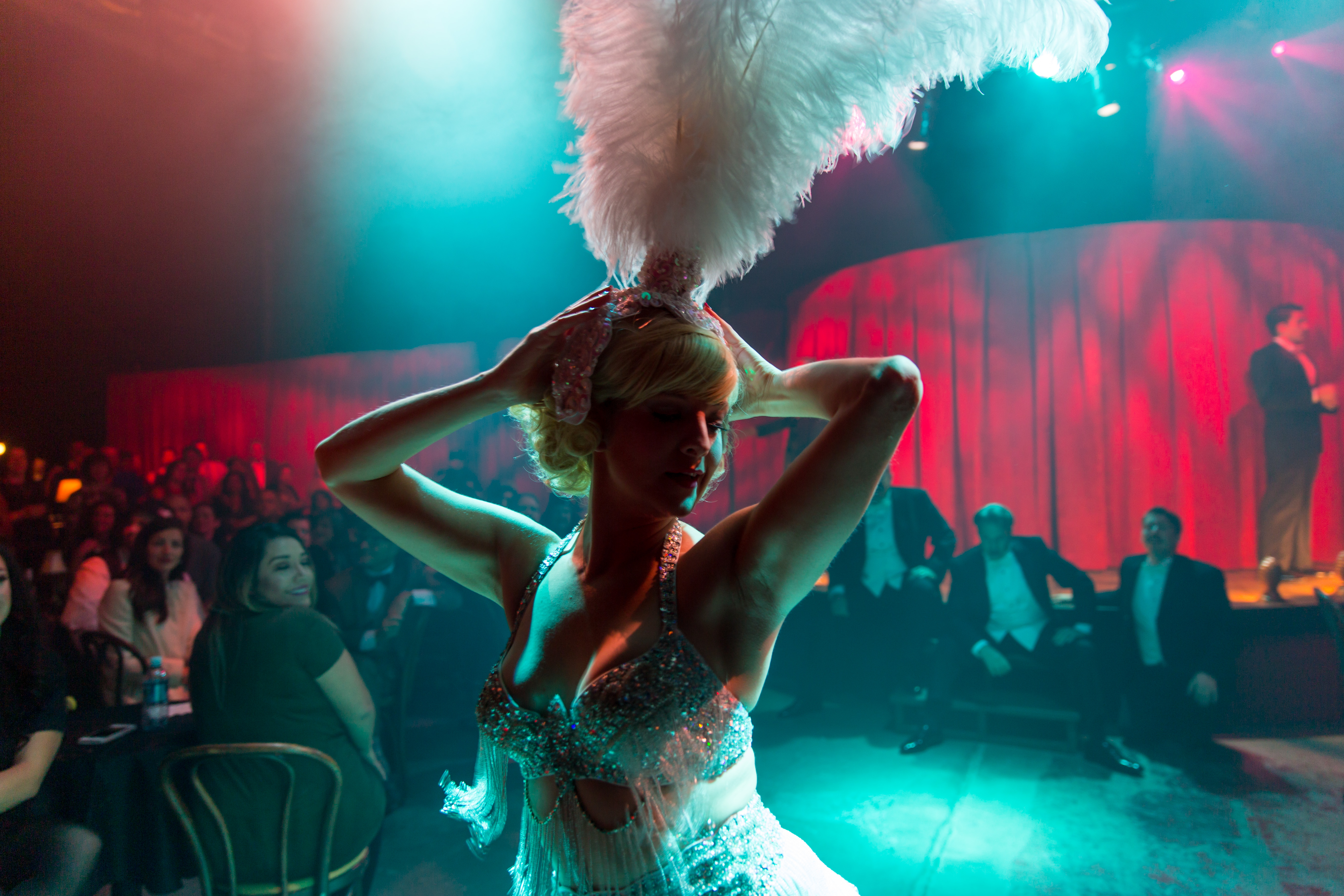 A woman is performing in a feather head dress while people look on. She is light from behind. 