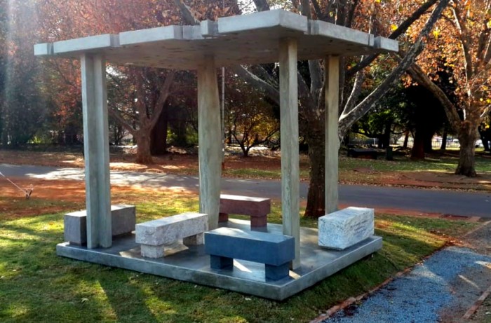 A memorial dedicated to South African trumpeter Hugh Masekela. Four columns hold up a flat concrete roof over five marble benches in an outdoor space surrounded by trees and grass..