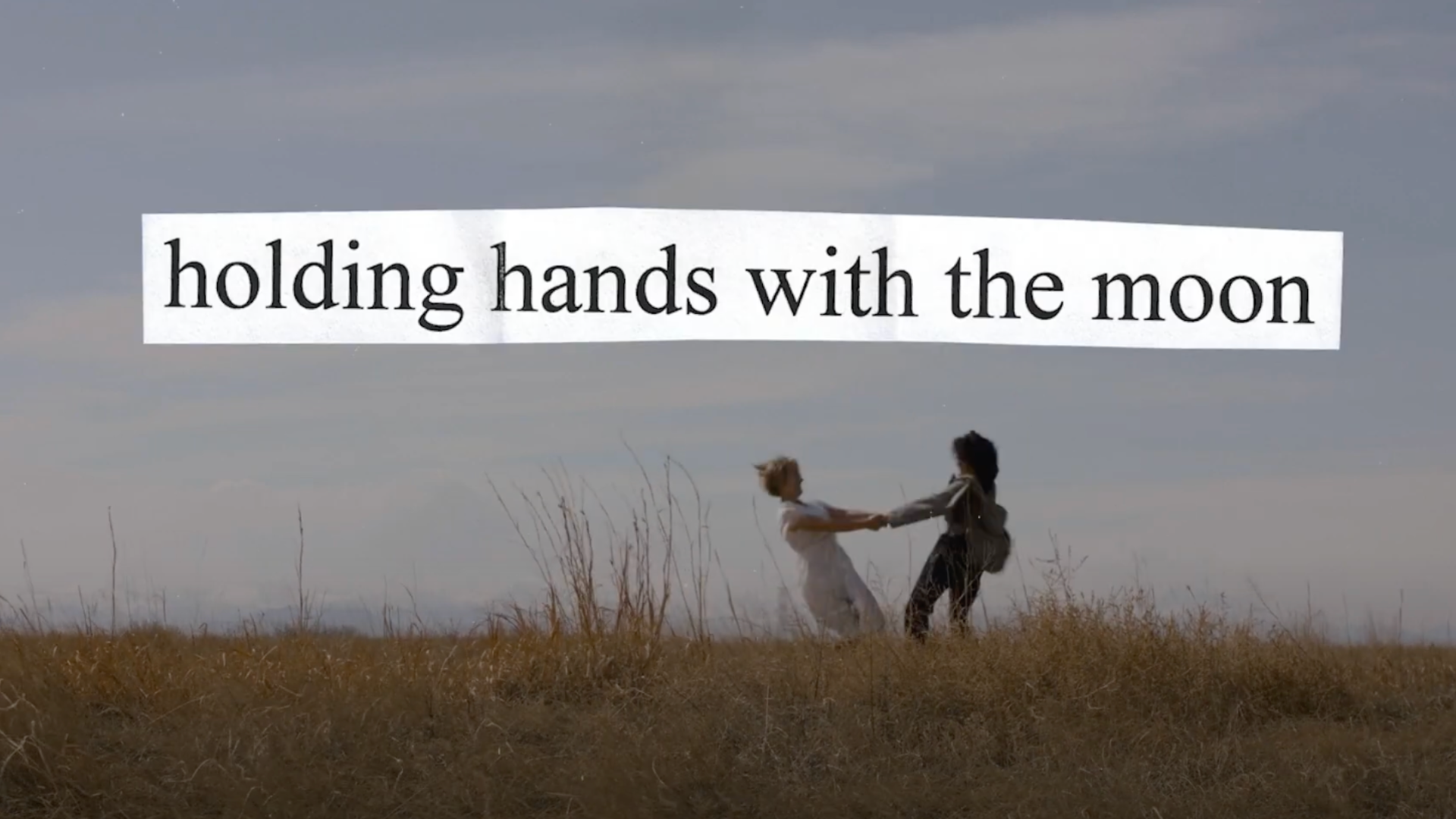 Film still featuring two people holding hands and spinning around in a circle. Text overlaid on the still image reads, "holding hands with the moon."