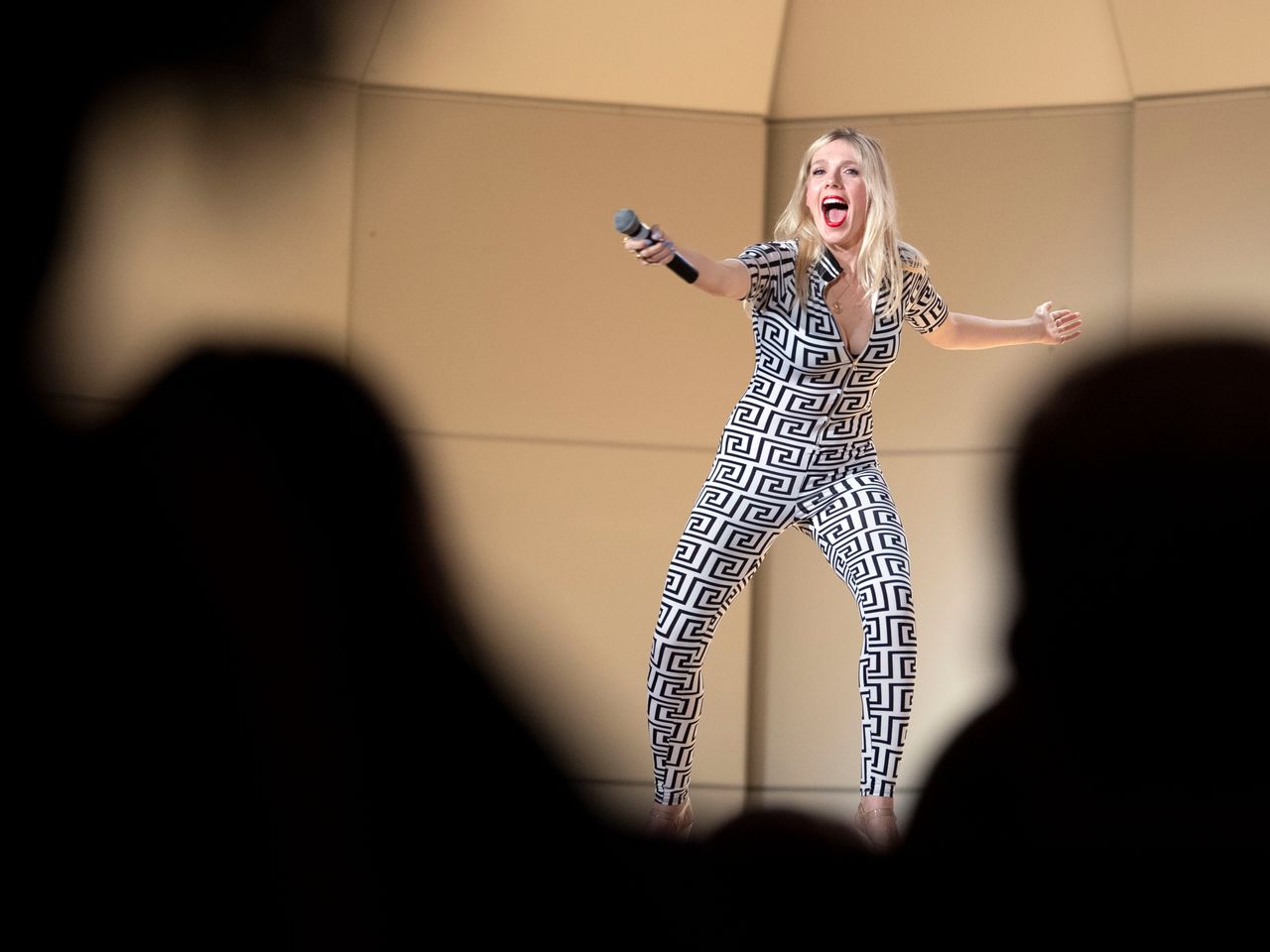 A blonde woman stands on stage in a black and white greco patterned body suit. She has her hand outstretched pointing a microphone towards the crowd. 
