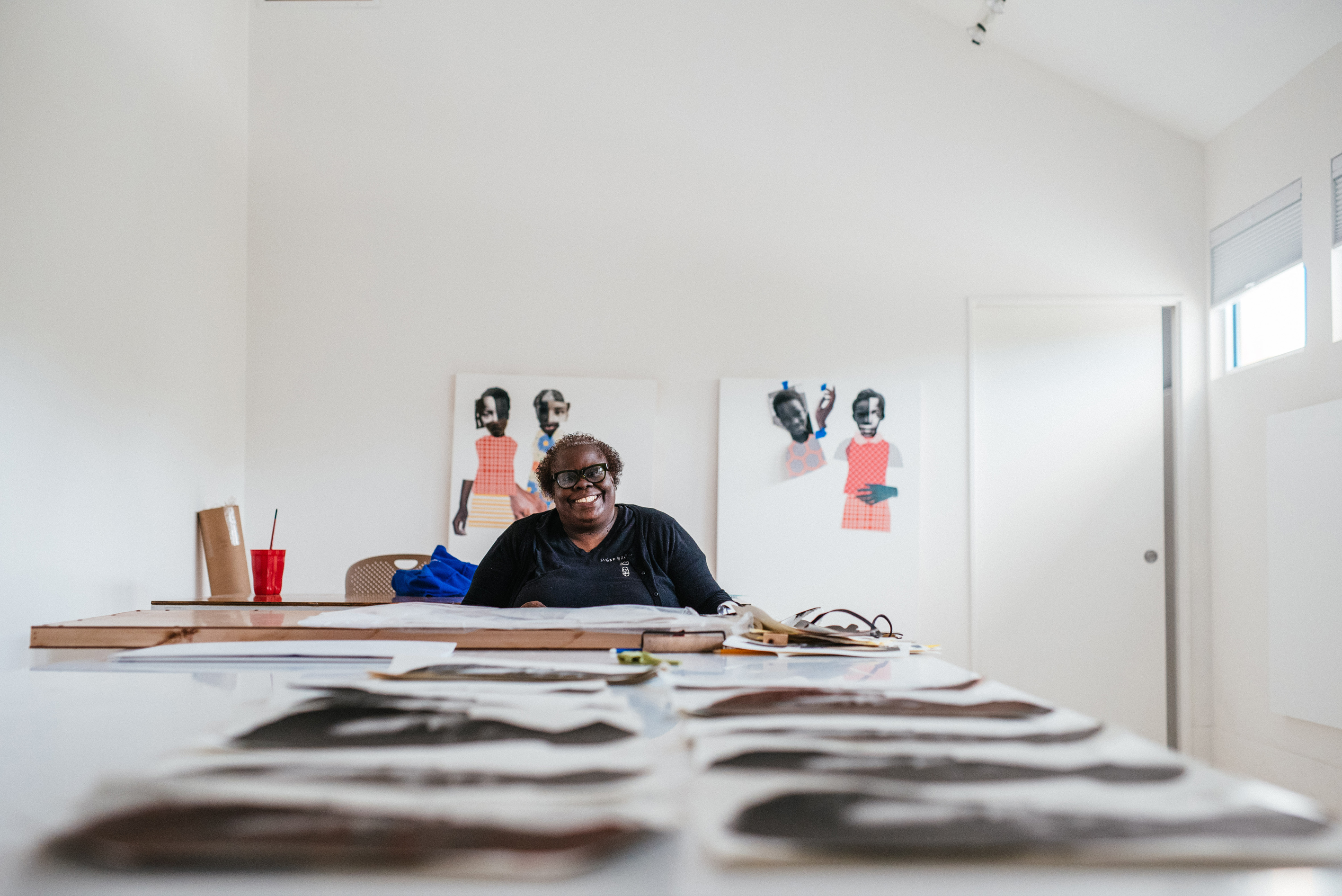 Artist Deborah Roberts sitting at a long table in a studio. She is sitting in the center of the frame, but is far away from the camera. Laid out on the table are, presumably,  in process works. Behind her, hung up on a white wall, are two in-process collage works of children. She is wearing a black shirt, angular glasses with black frames, and has a big, beautiful smile on her face. 