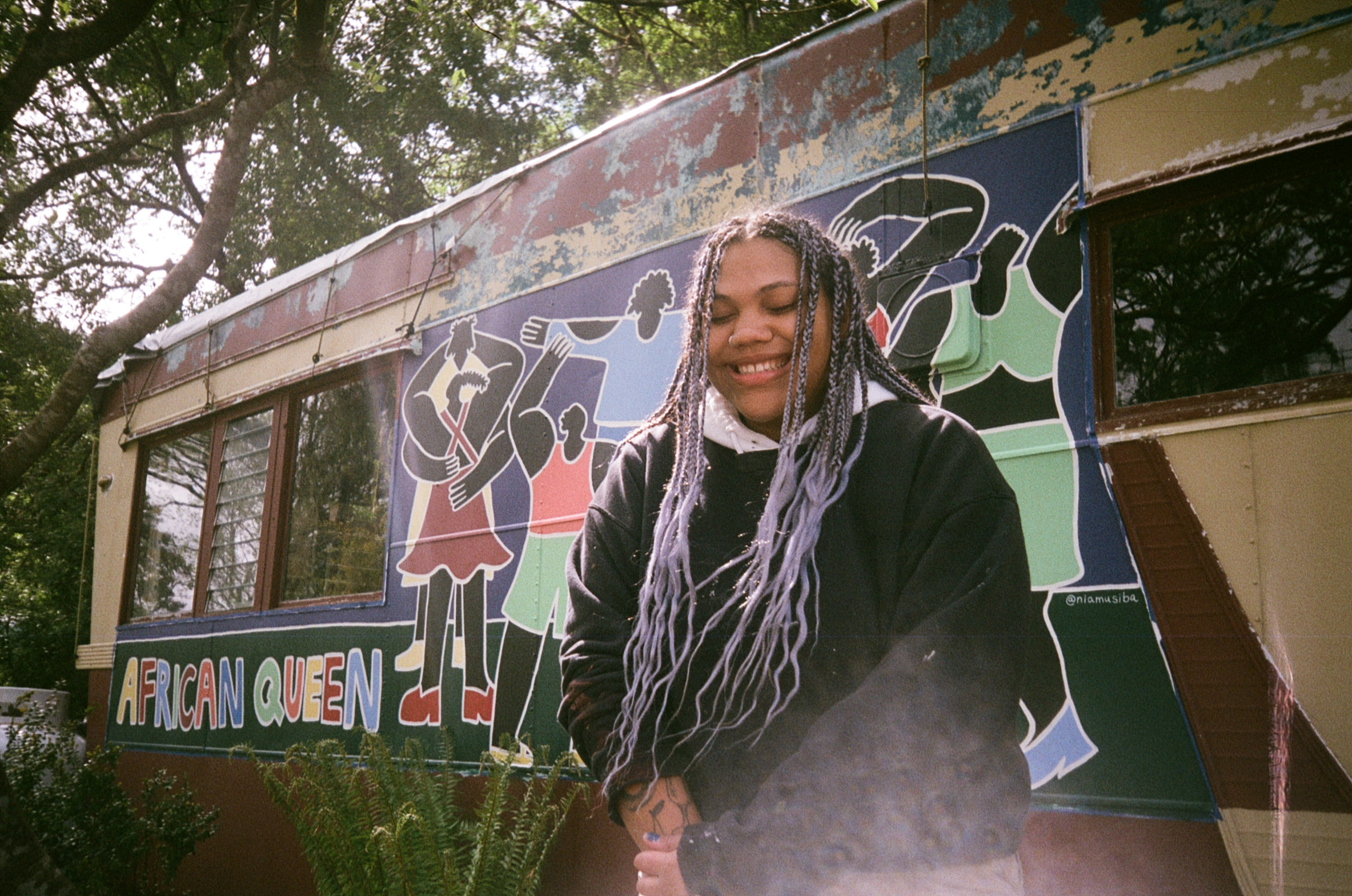 Artist Nia Musiba standing outside, in front of a mural with figures dancing and hugging. Text on the mural reads, “African Queen” and the mural itself is nestled in some trees. Nia is wearing a black hoodie and is sporting purple braids. Her eyes are closed and her smile is beaming.