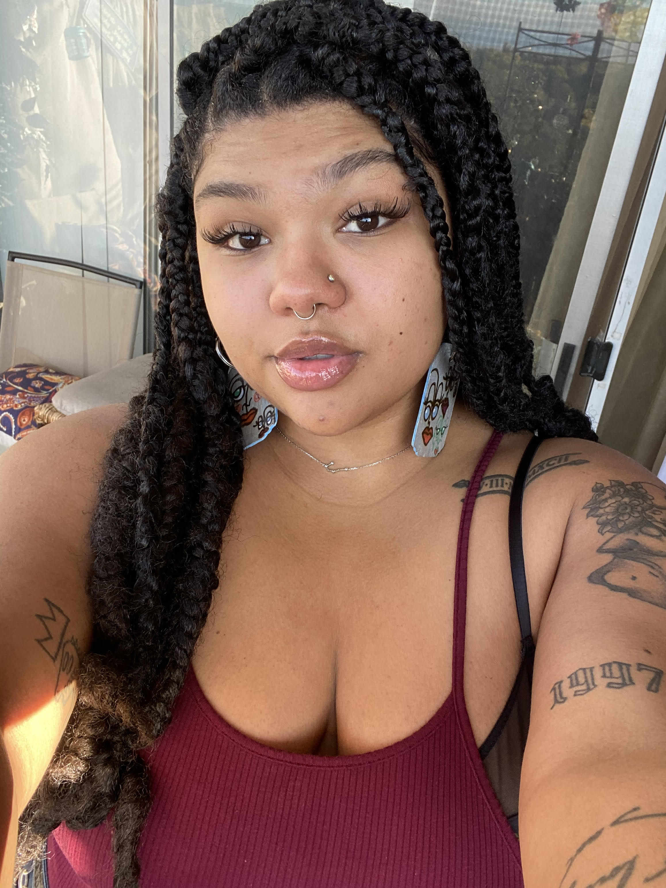 A selfie of Christina, wearing a burgundy tank top, which reveals the tattoos on her arms. Her lips are pierced, her hair is braided, and she has large earrings on that have faces on them. 