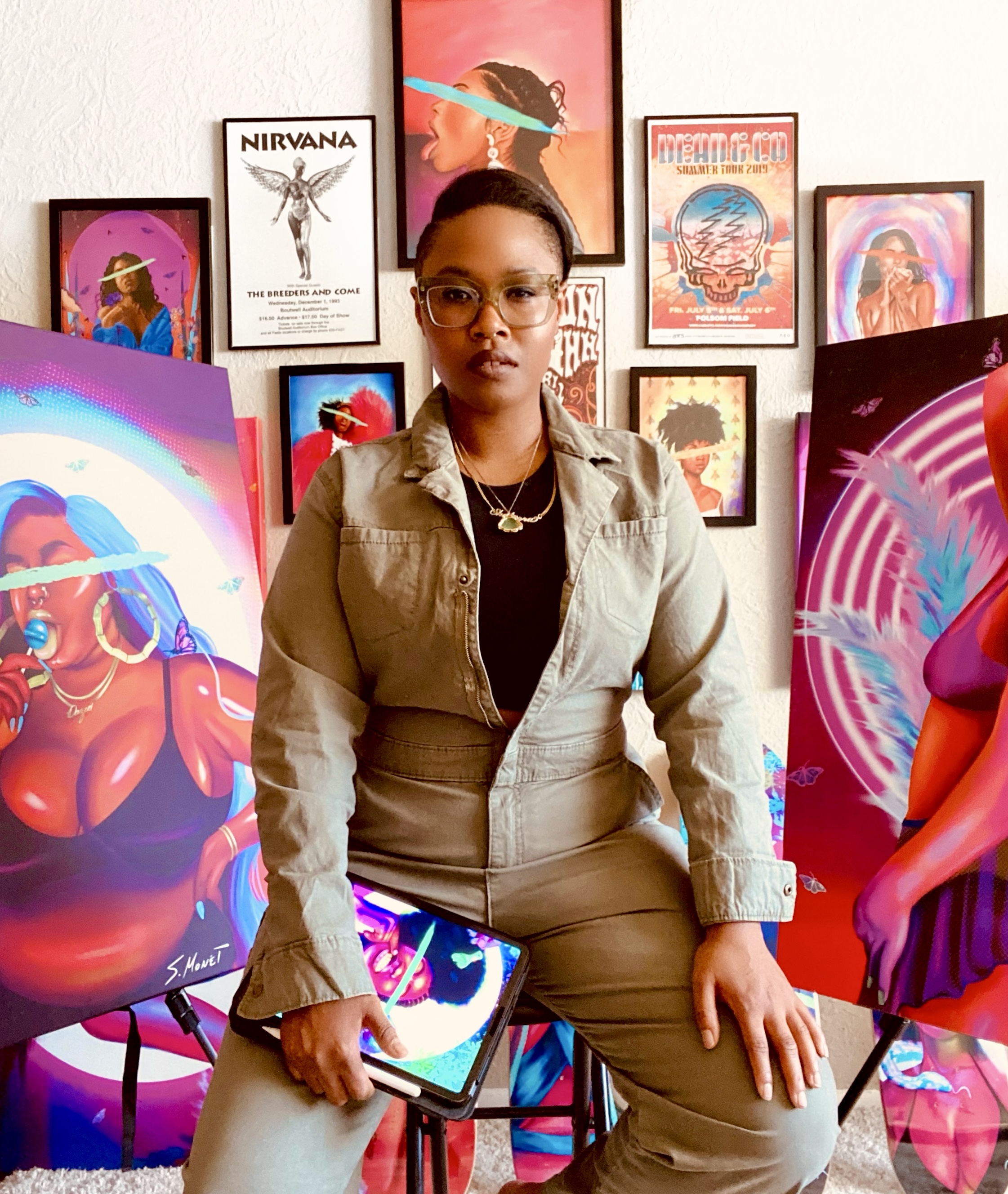 Contemporary artist S. Monét sitting on a stool in her art studio. She is sporting a jumpsuit, black t-shirt, and glasses, and has a serious look on her face. Behind her is colorful artwork hung on the wall and large canvases. 