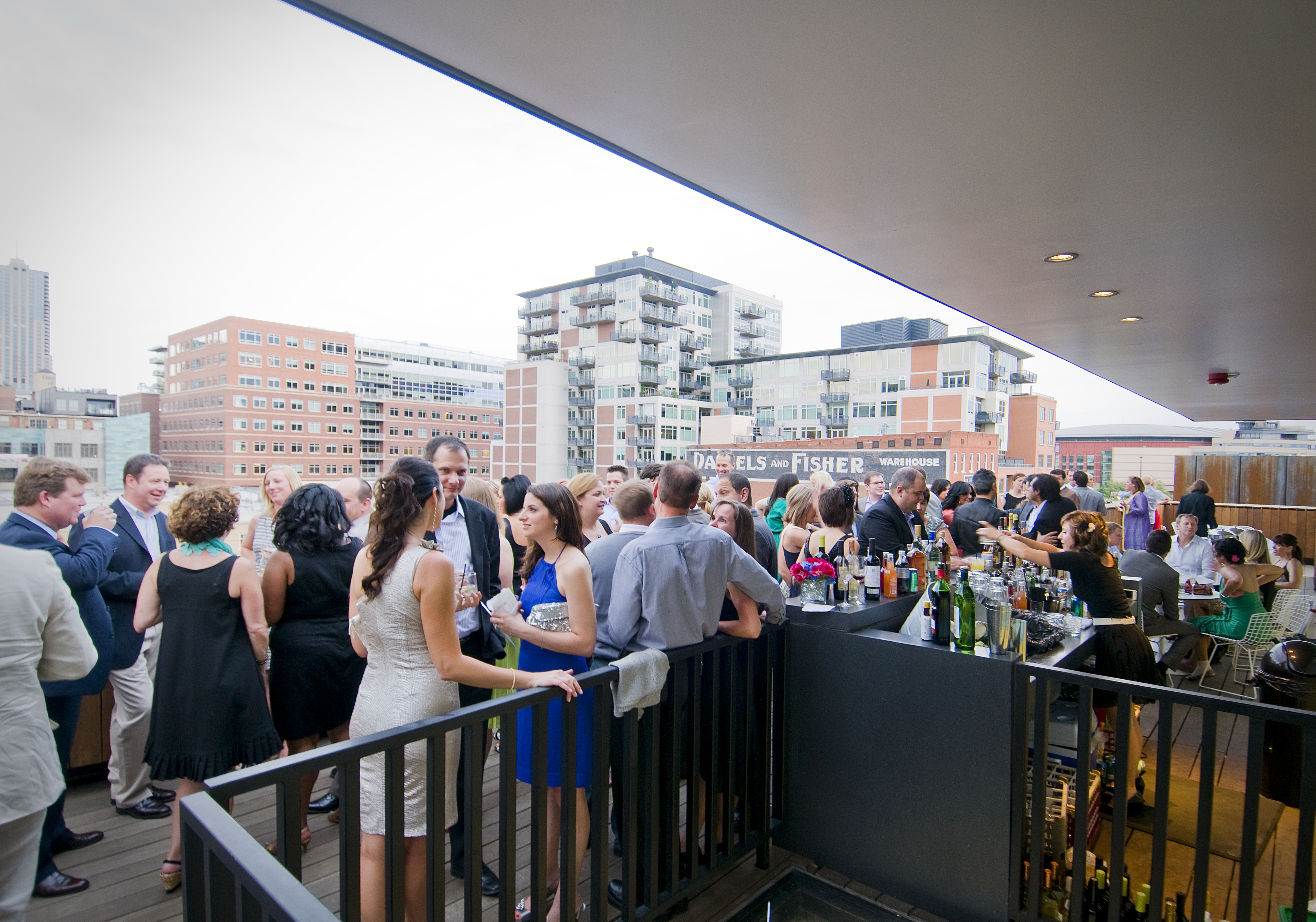Dozens of people gather on the rooftop surrounding an outdoor bar. They are wearing dresses and suits, chatting, smiling, and enjoying beverages. 