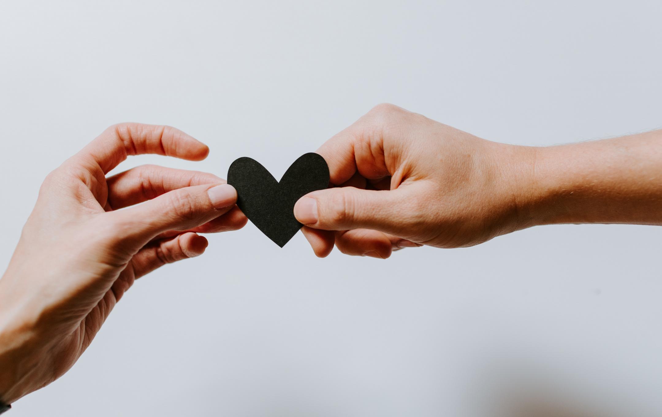 Two hands are gently holding a black paper heart in front of a white background