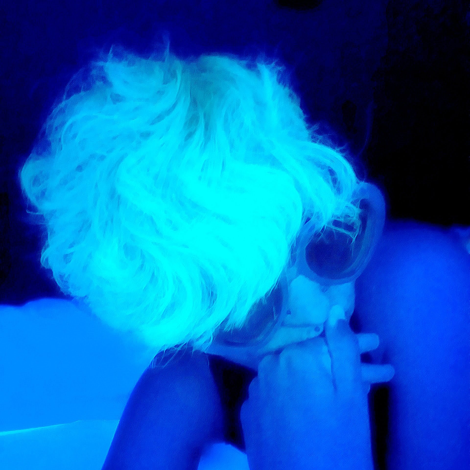 [Image description: Photo of Diamanto Sala. There is a heavy blue filter on the photo of Diamanto in a pensive pose. They are wearing large sunglasses and their short wavy hair is glowing in the blue light.]