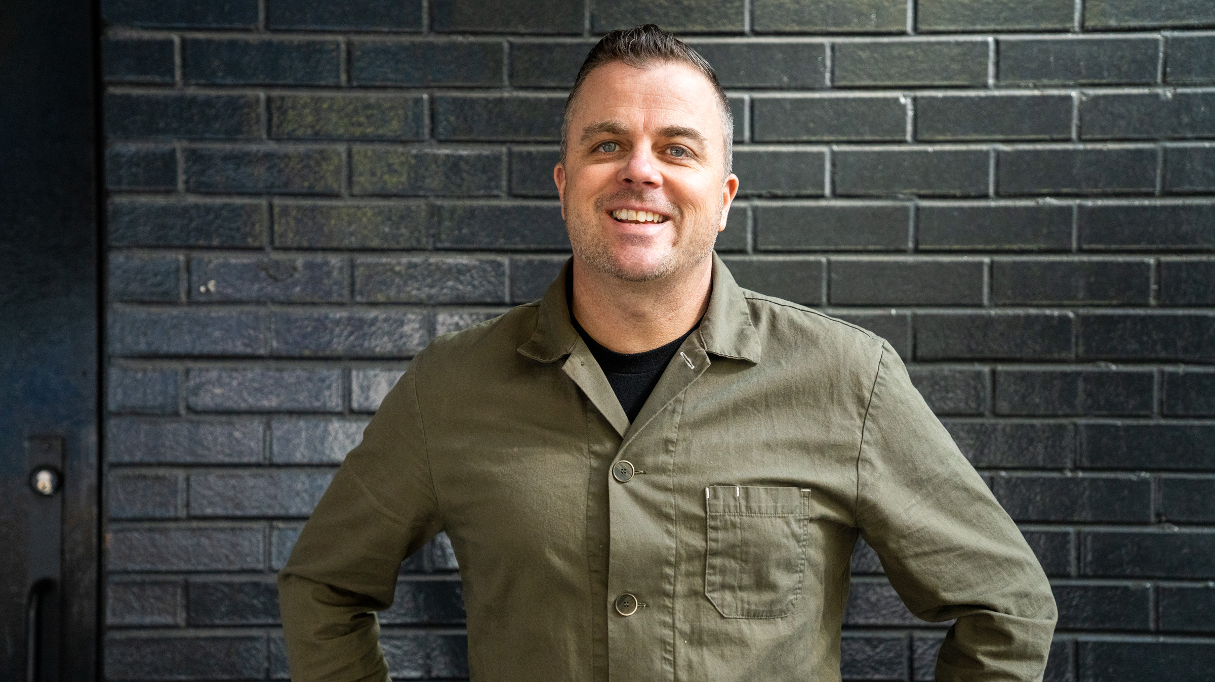 [Image description: Chef Kelly Whitaker stands in front of a gray brick wall with his hands on his hips. He wears an army green collared button down shirt and his salt and pepper hair in a short crop.]
